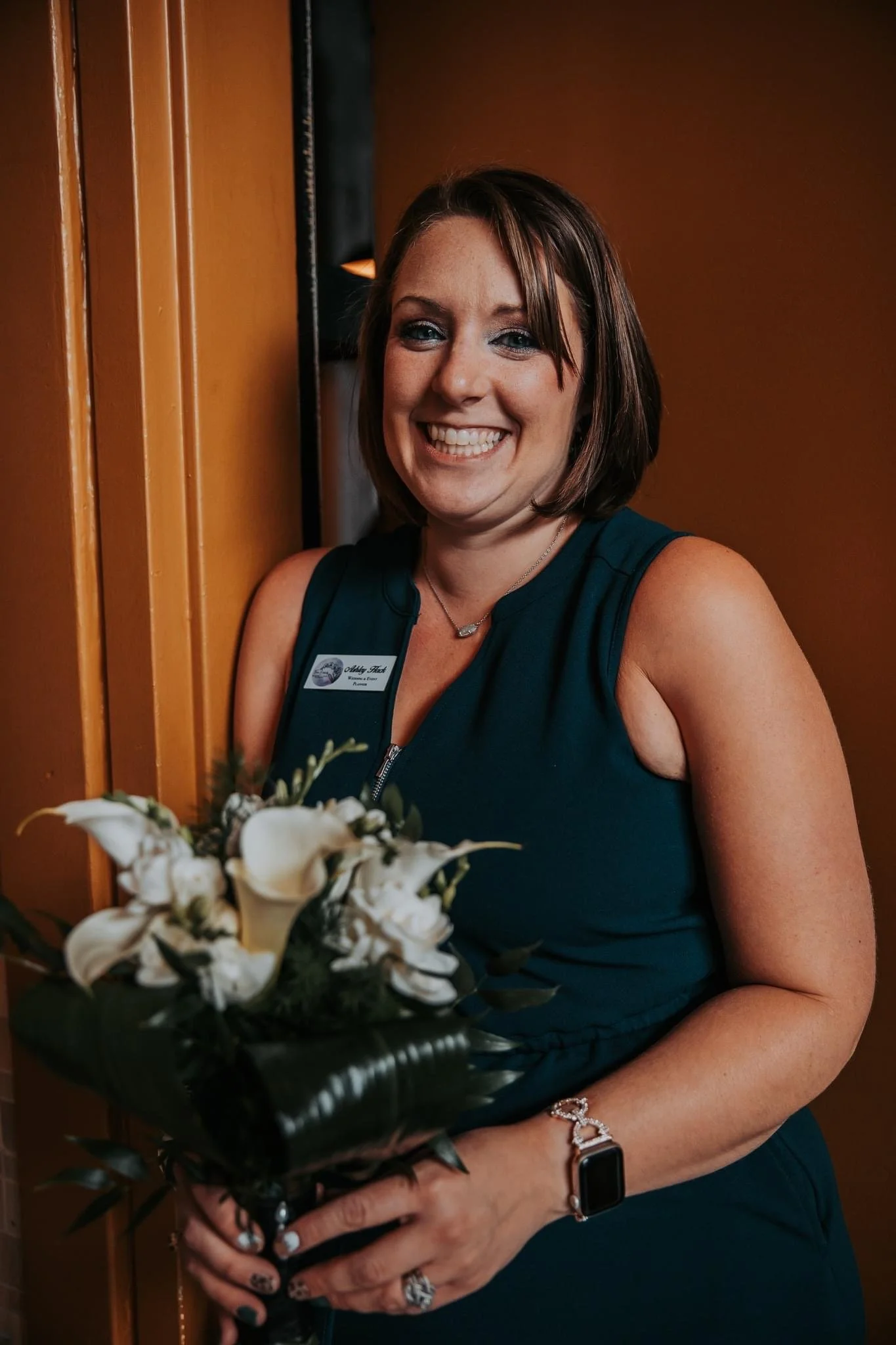 Smiling woman holding flower bouquet, wearing a teal dress and a name tag, standing against a yellow wall.