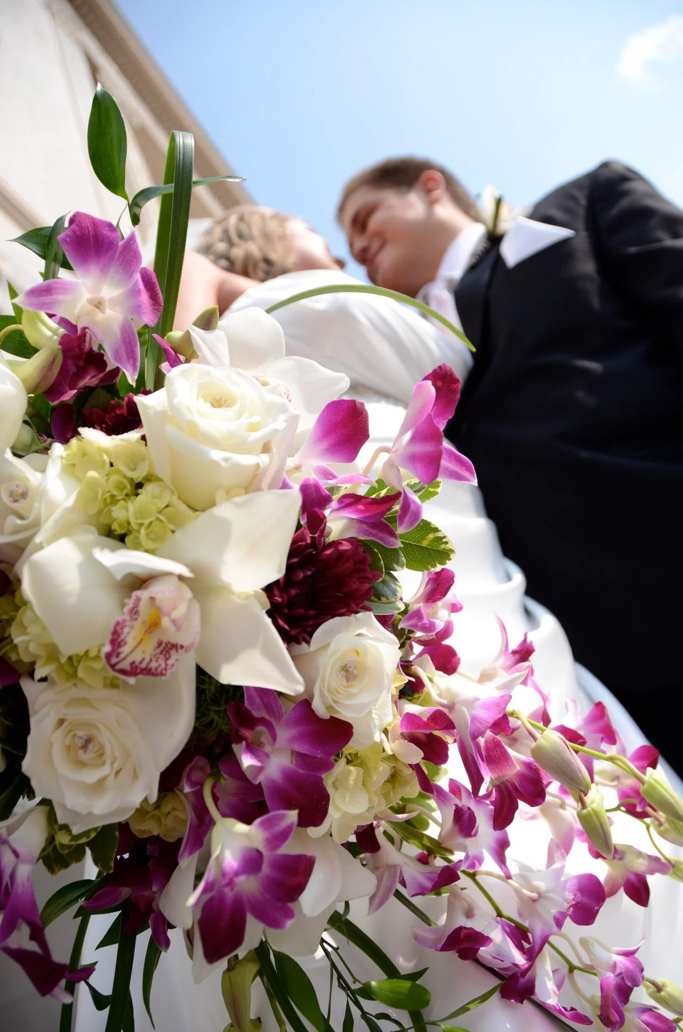 Close-up of a wedding bouquet with white roses, pink and purple flowers, and greenery, with a bride and groom in the background.