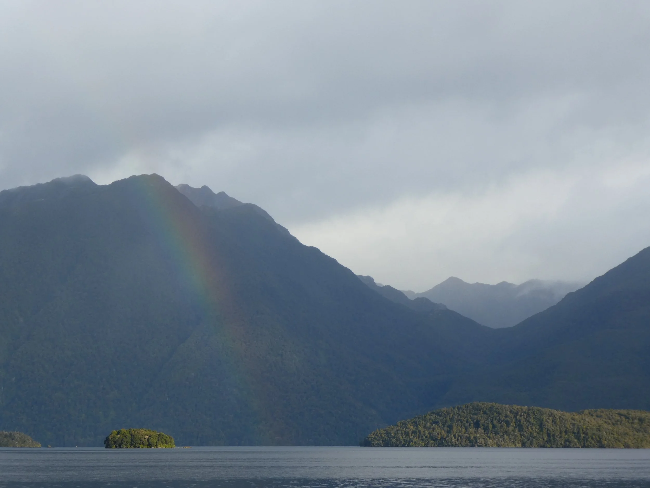 Mountain landscape with a rainbow over a large body of water and forested islands.