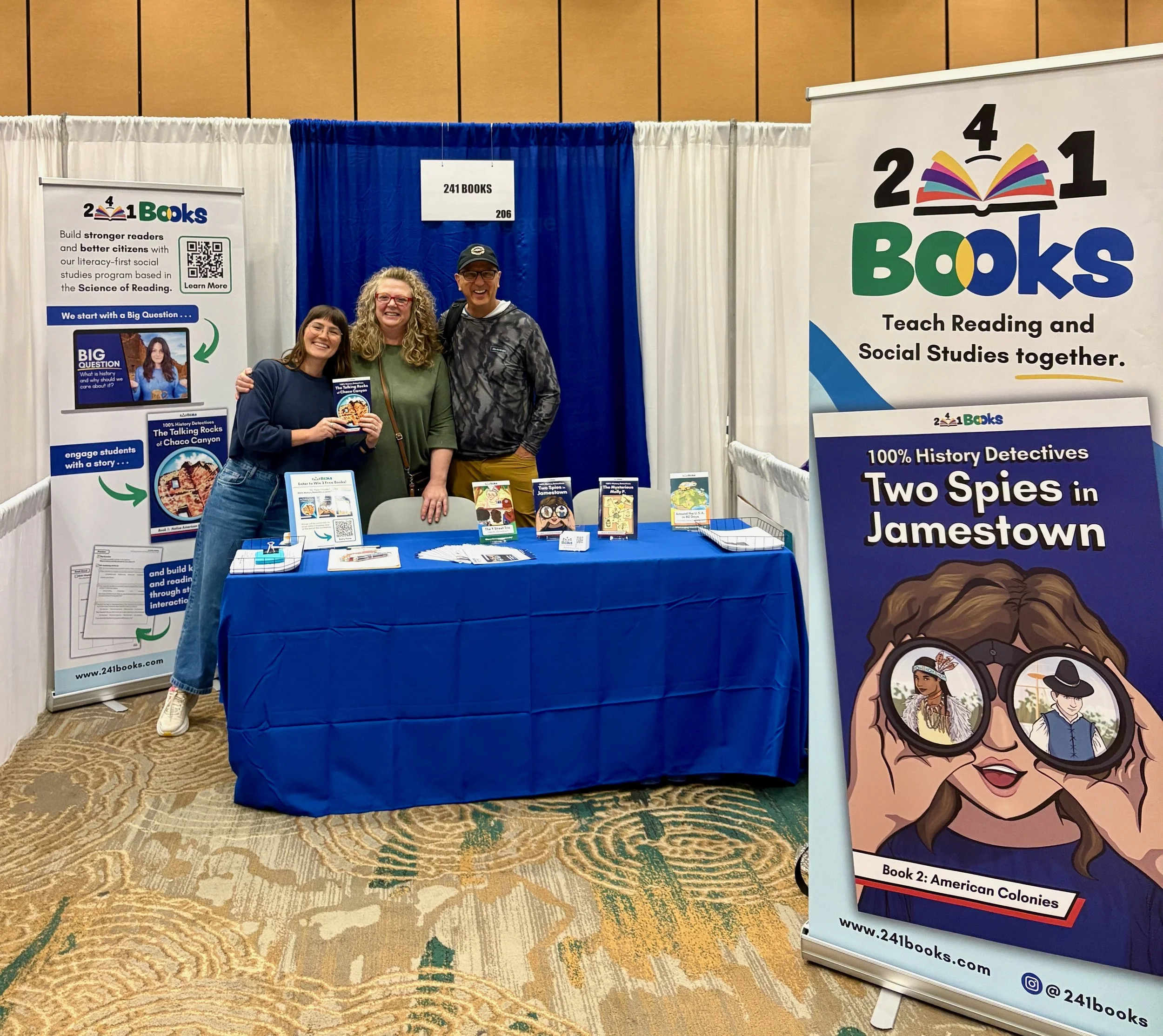 The founders of 241 Books stand smiling at an educational conference. They hold a history book titled "Two Spies in Jamestown", which is part of a nine book series. Their shirts read "Stronger Readers. Better Citizens."