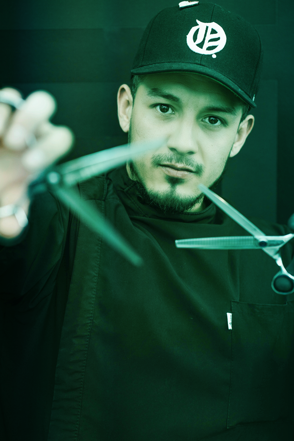 Young man with a beard wearing a black jacket and a cap with a logo, holding hair cutting scissors and clippers, looking at the camera. Background is dark.