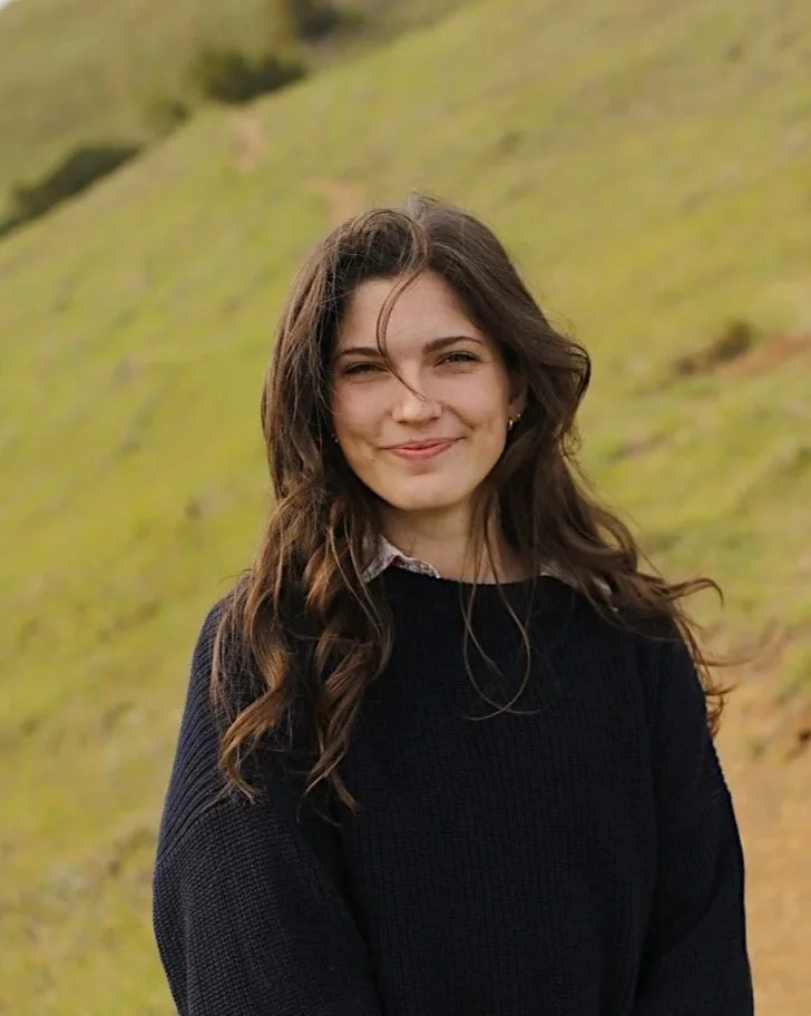 Young woman with long wavy brown hair standing outdoors on a grassy hillside, smiling at the camera, wearing a black sweater.