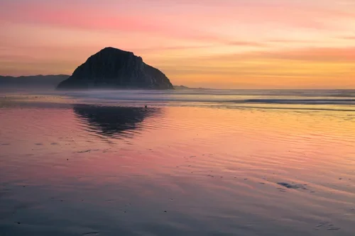 Sunset over a calm beach with a large rock formation in the distance and pastel-colored sky reflected in the water.