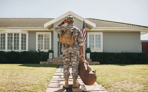 A service member saluting in front of a suburban home displaying an American flag.