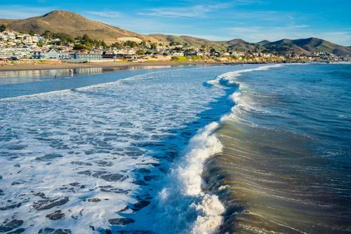 Waves crashing on a sandy beach with a beachfront town and hills in the background.