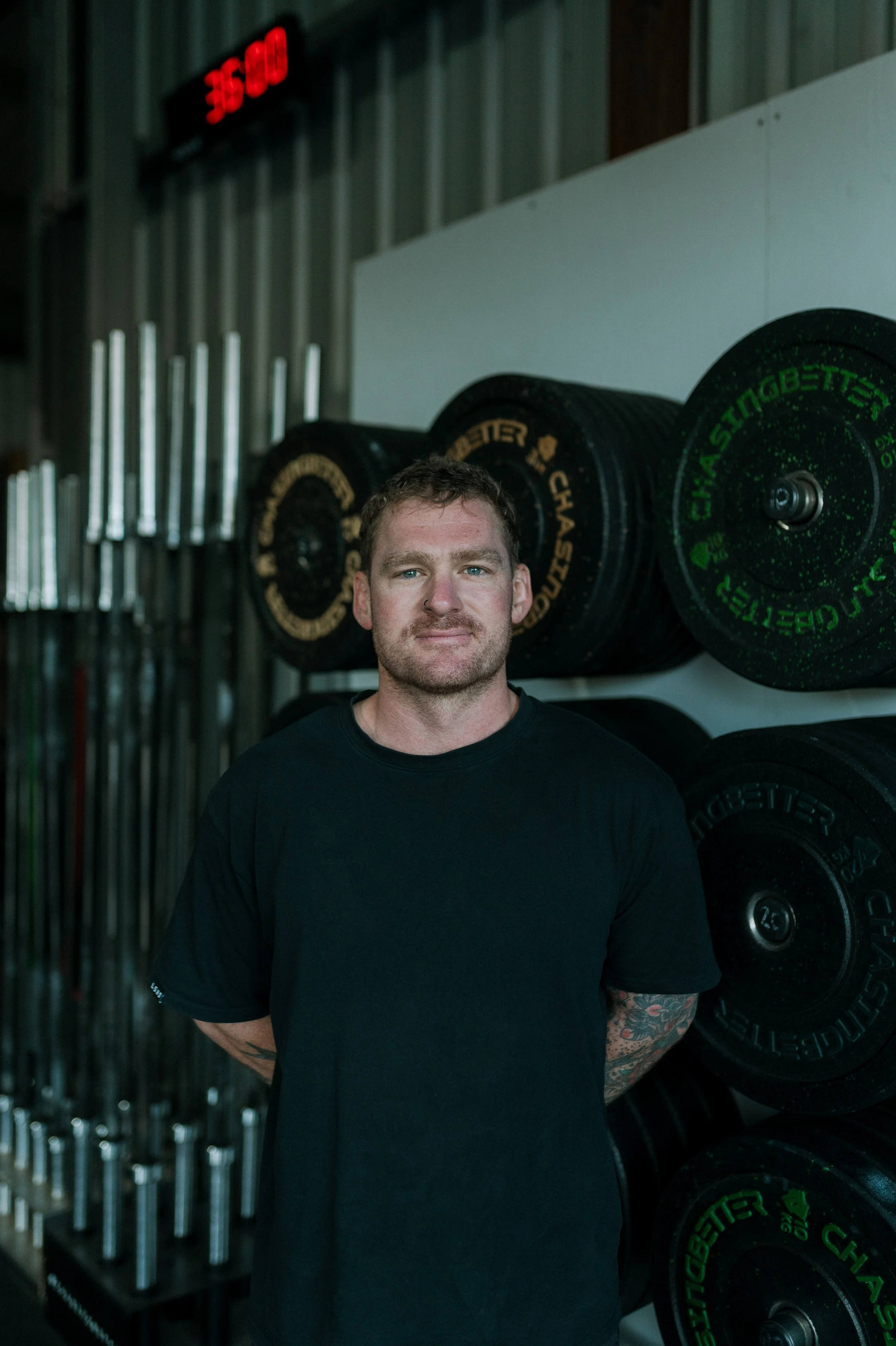 A man with short, curly hair and a beard stands in front of black and green weight plates on a rack in a gym, with a steel wall and fluorescent lights in the background. The digital clock shows 3:50.