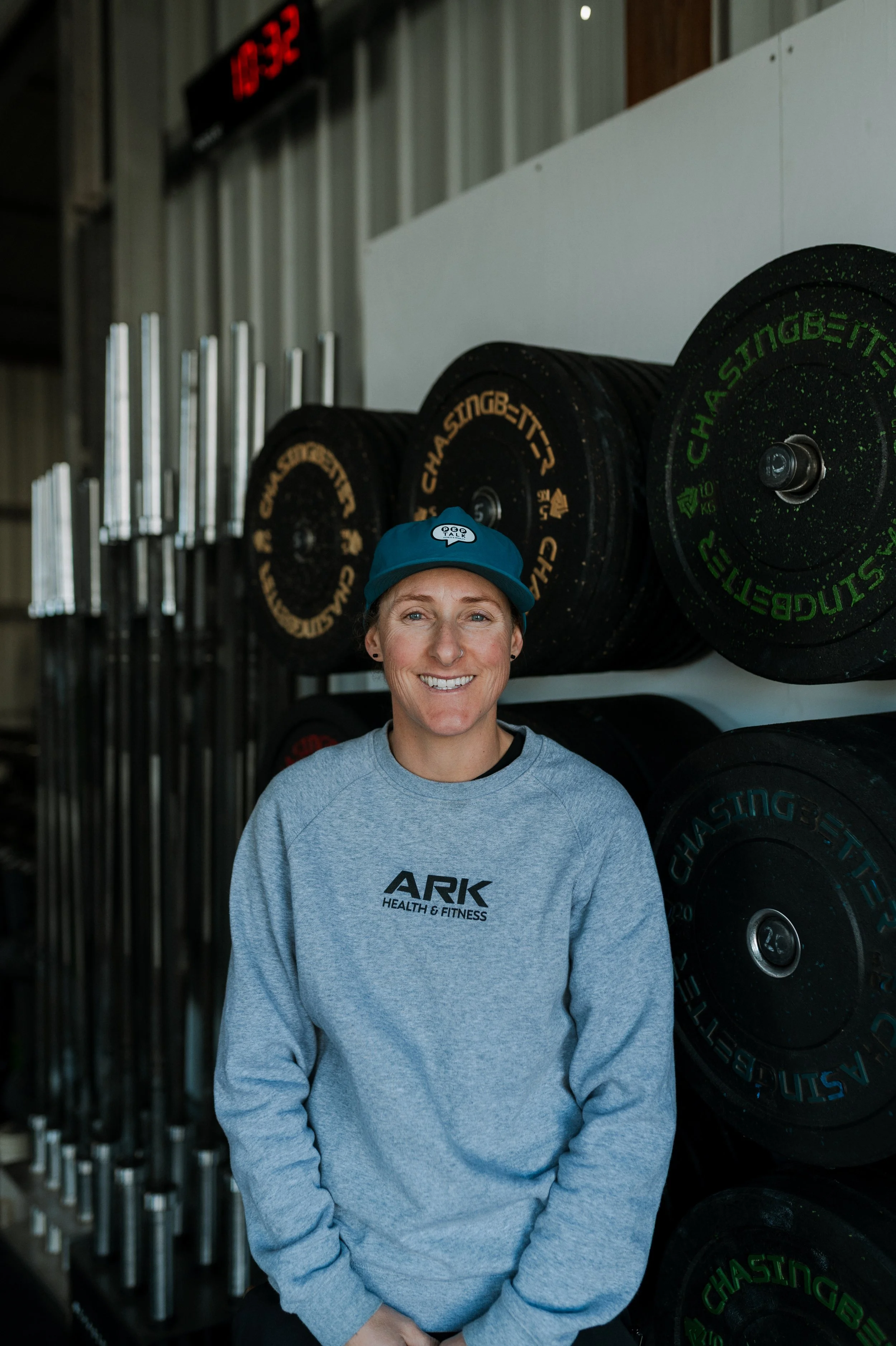 A smiling woman wearing a blue cap and a gray ARK Health & Fitness sweatshirt standing in a gym next to stacked black weight plates with green and yellow markings and vertical metal bars in the background. A red digital clock shows 16:32.