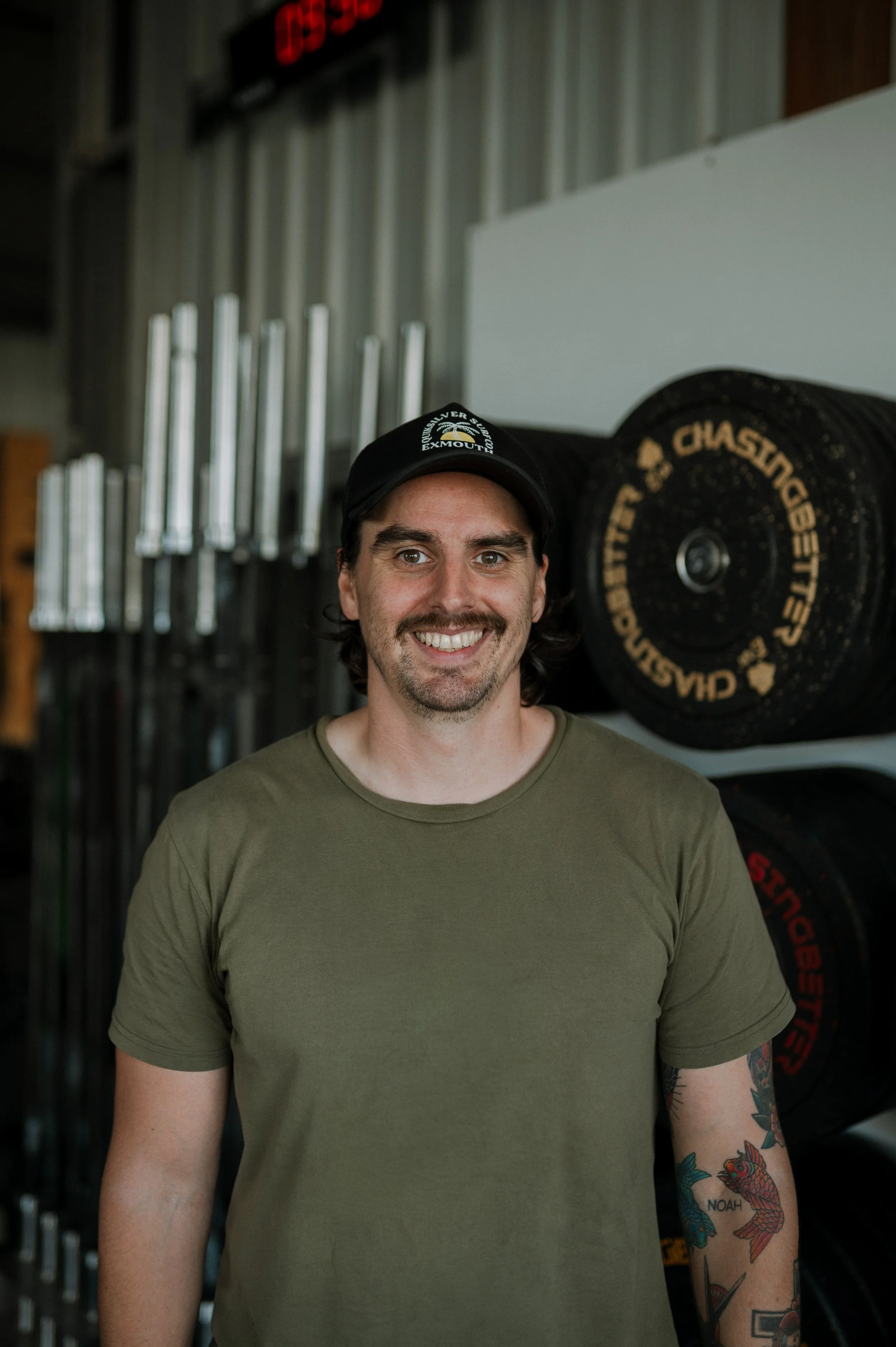 Smiling man with dark hair, wearing a black cap with white text, green t-shirt, and colorful tattoos on his left arm, standing in front of a wall with weightlifting equipment in a gym.