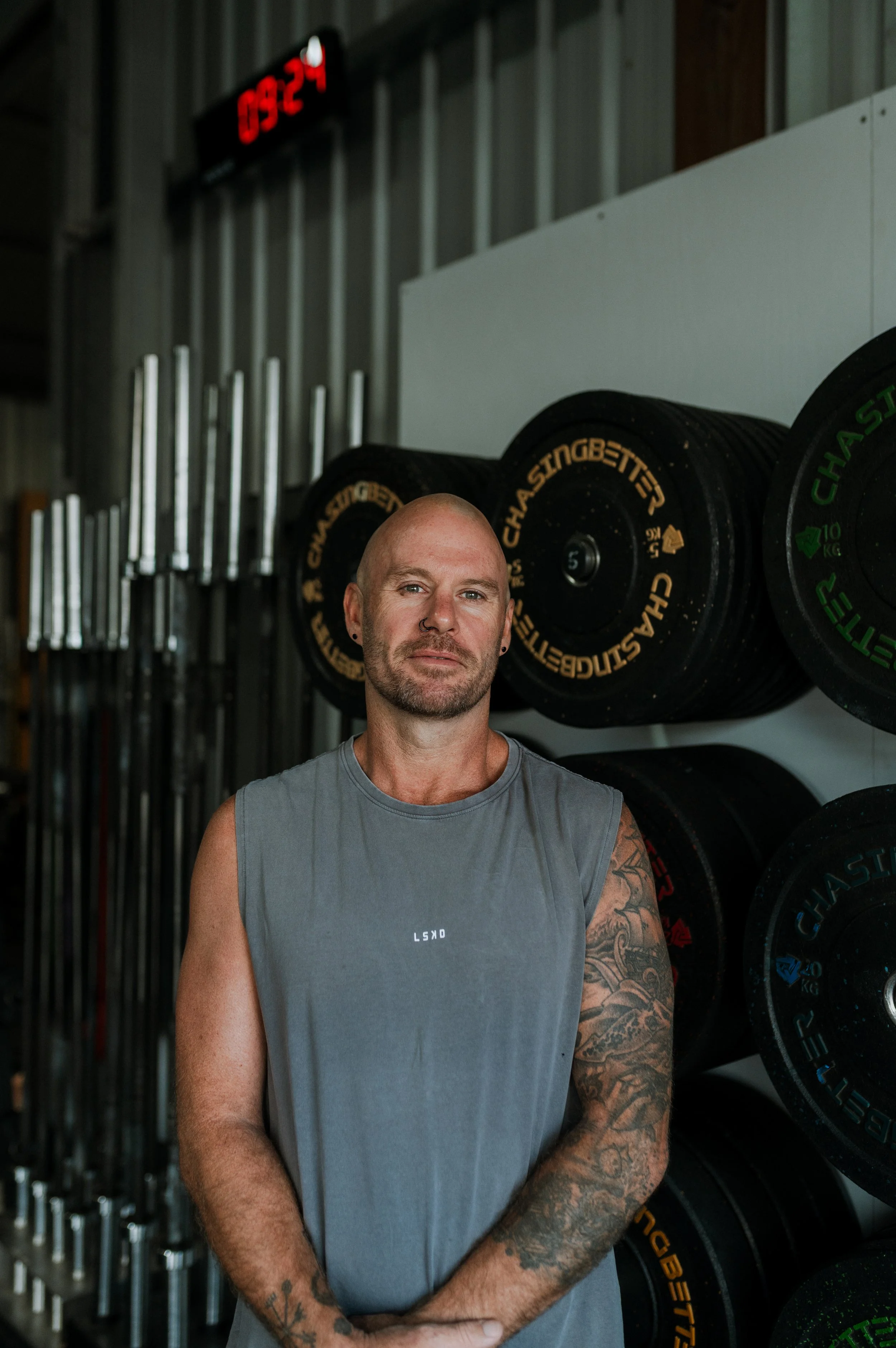 Man with tattoos wearing sleeveless gray shirt standing in gym with weighted plates and barbells in background.