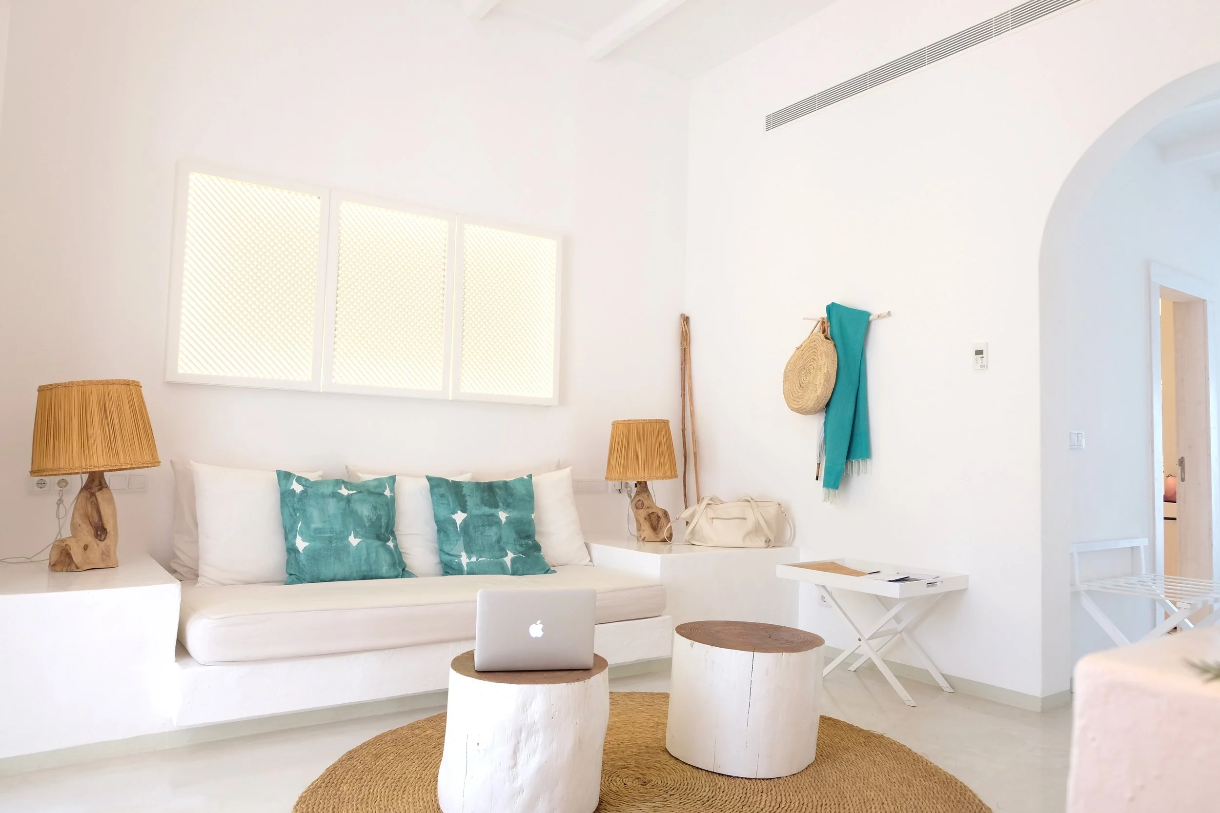 Minimalist white living room with white sofa, teal cushions, two wooden lamps, a MacBook laptop on a white wooden coffee table, wicker bags, and wall decorations in a bright, airy space.