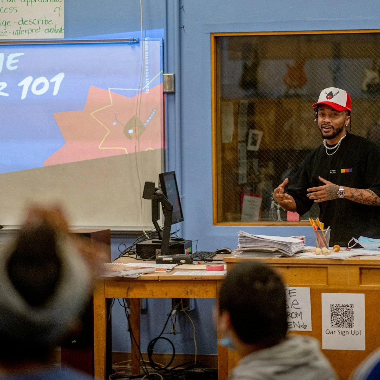 A man wearing a black shirt, red and white baseball cap, and jewelry is giving a presentation in a classroom. There is a computer, piles of papers, and supplies on a desk. Students are watching, and a whiteboard with a projected slide and a window with a wire mesh are in the background.