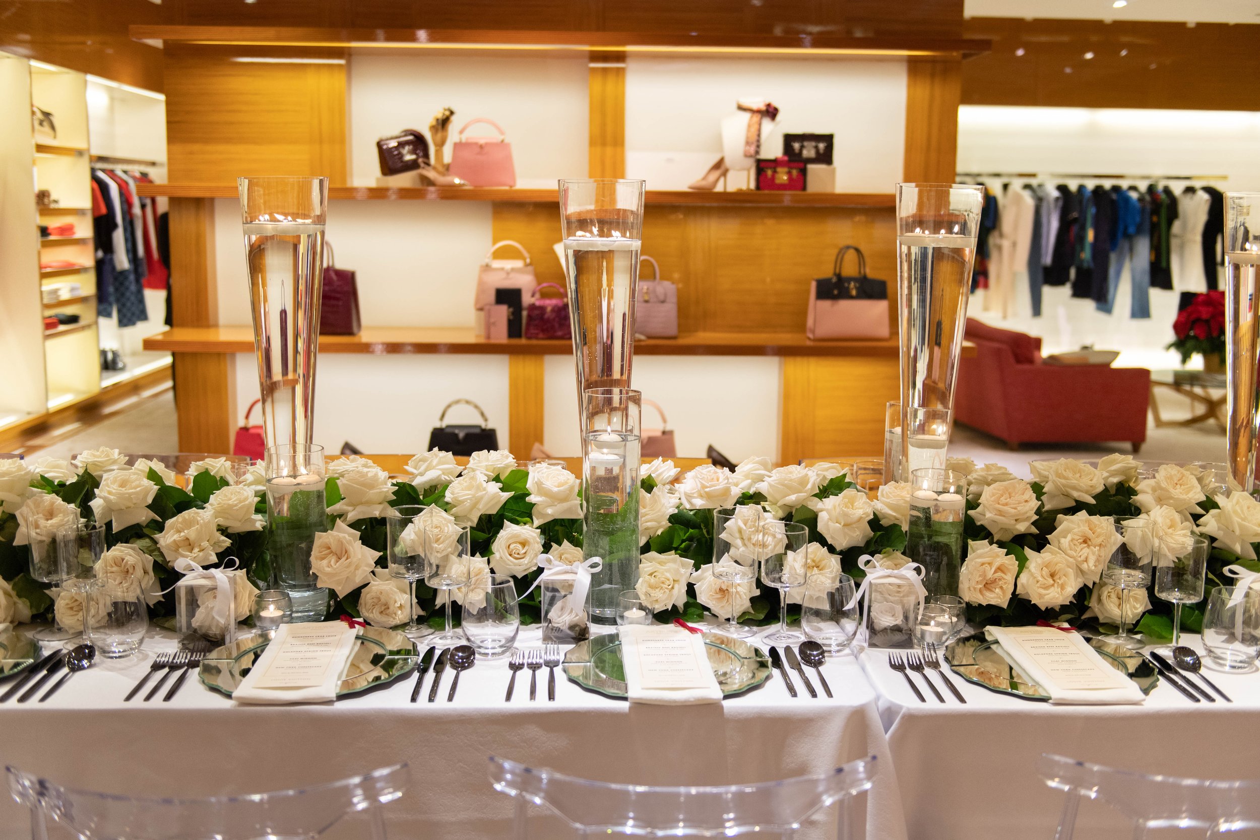 Elegant dining table setup with white roses, tall glass vases with floating candles, and place settings including plates, silverware, and stemware, in a store with handbags and clothing displays in the background.