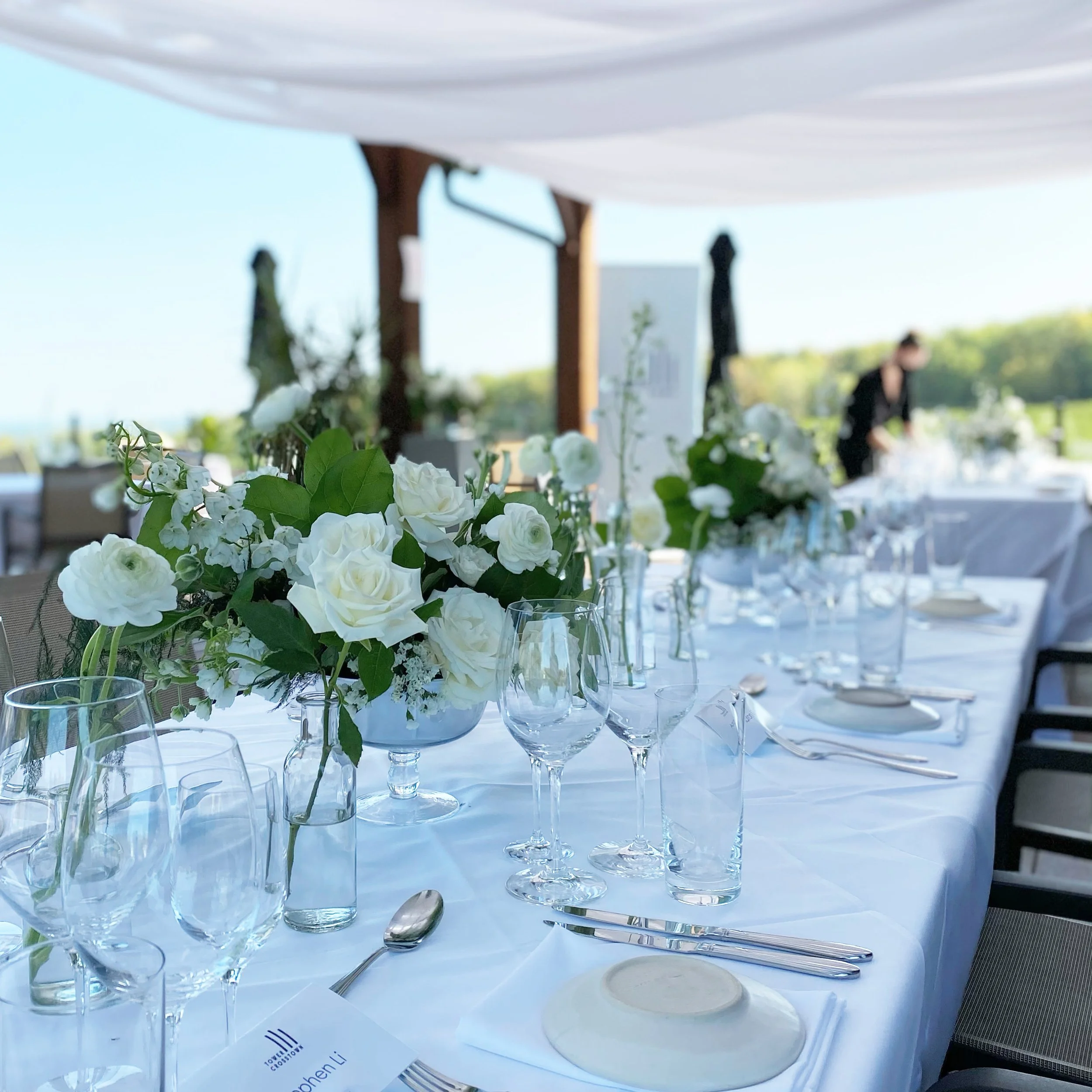 Elegant outdoor wedding reception table with white floral centerpieces, glassware, and fine tableware, set under a canopy with green landscape in the background.