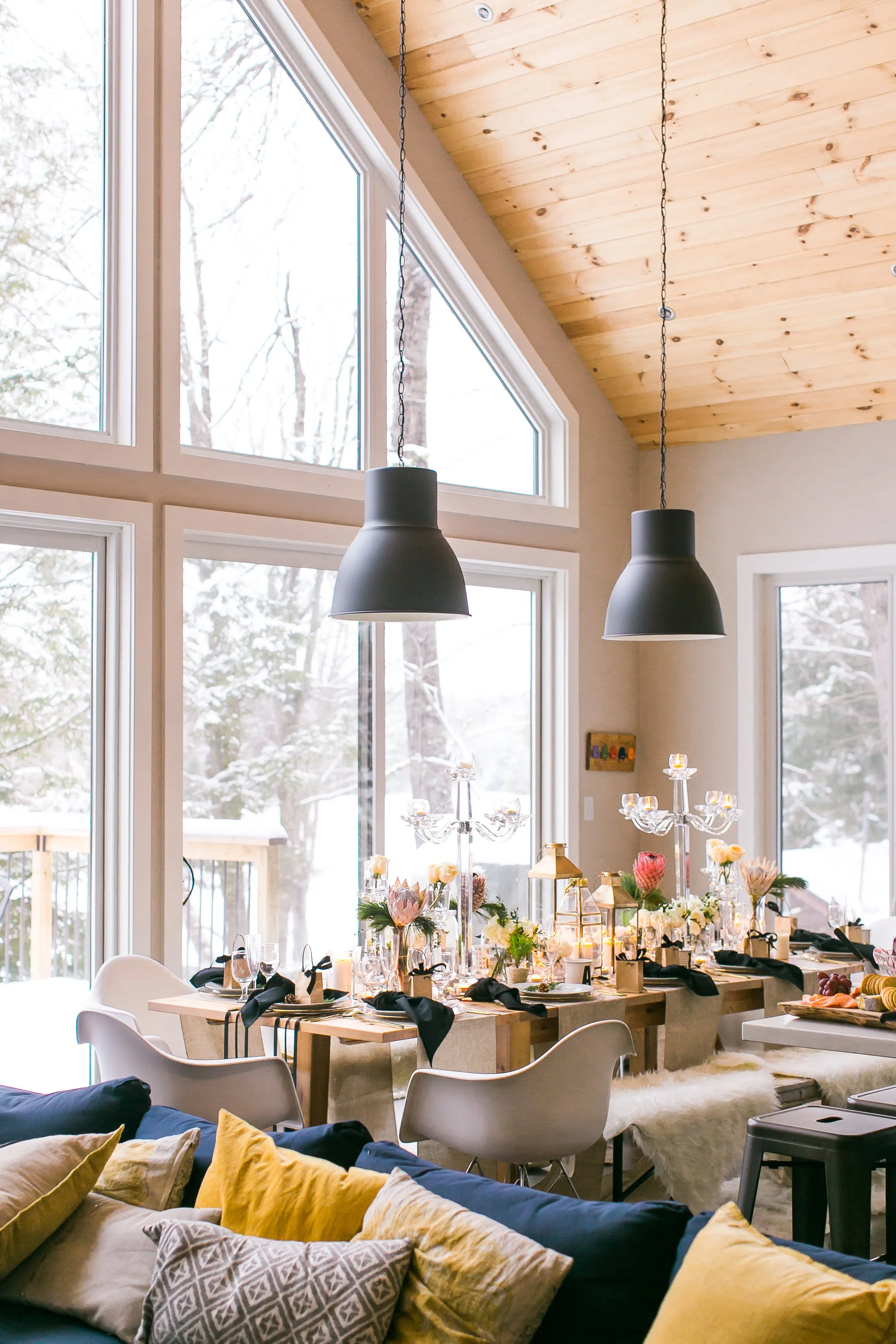 Interior of a dining area with large windows showing a snowy outdoor scene, decorated for a meal with flowers, candles, and tableware.