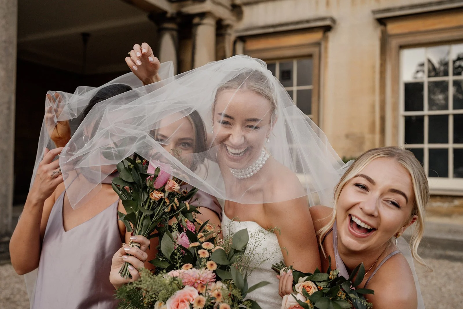 A bride and her bridesmaids celebrate, smiling and holding bouquets, with the bride’s veil draped over them all during a wedding.