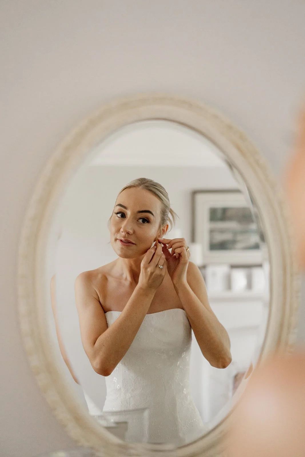 A woman in a white wedding dress is putting on earrings while looking into an oval mirror.