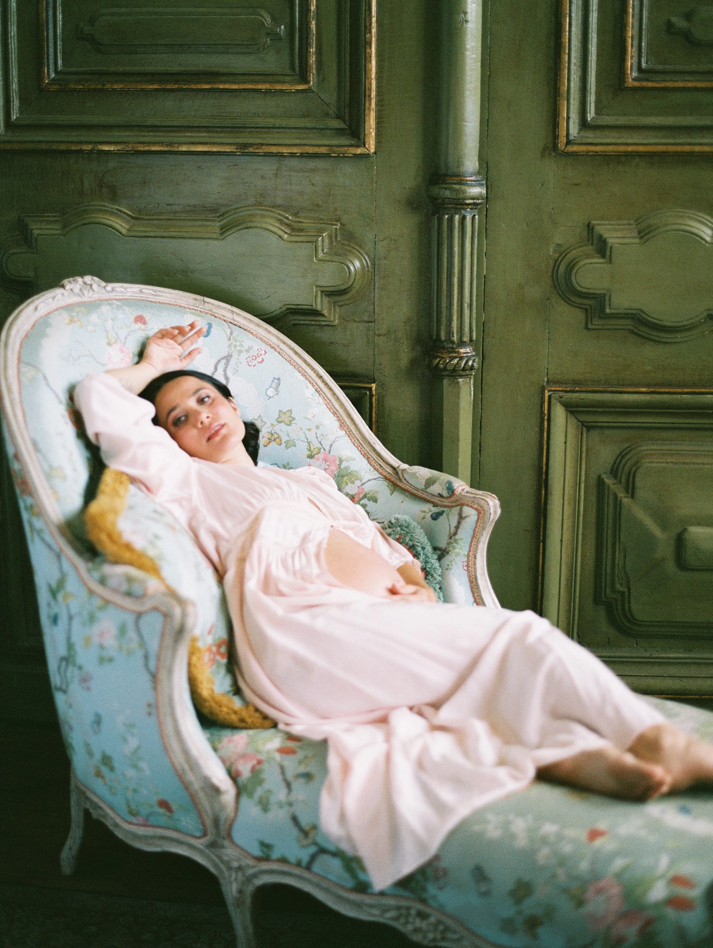 A pregnant woman reclines on a​n antique​ French chaise. She wears a pale pink robe ​and her round stomach​ is exposed​. She gazes directly at the camera. Behind her is a green​, antique do​or.