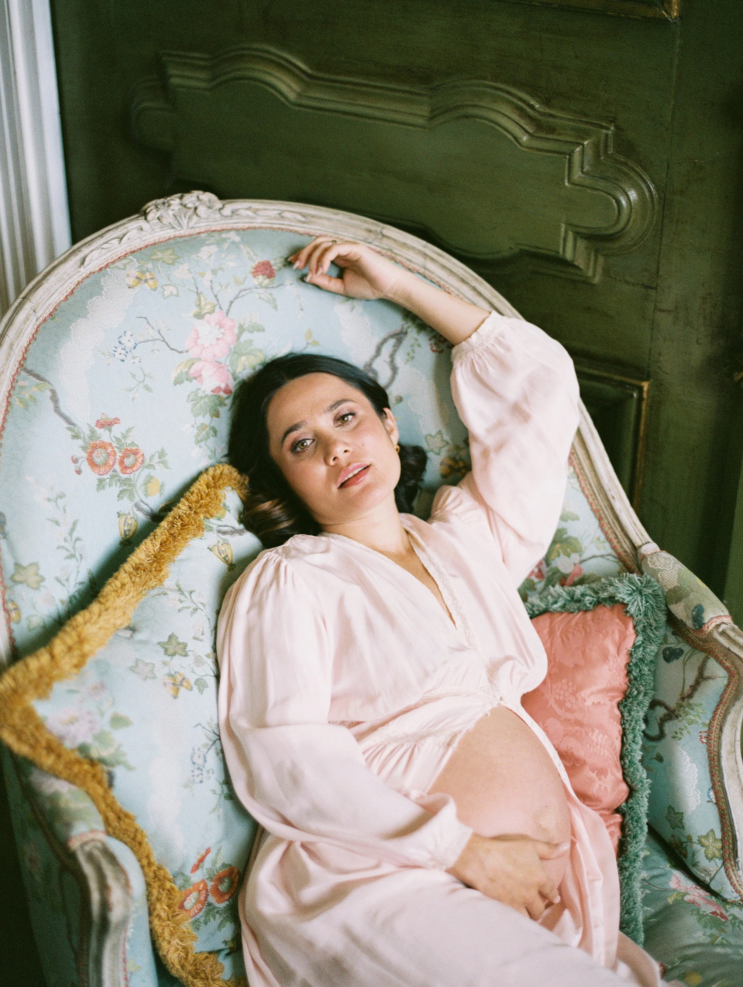 A pregnant woman poses on a​n antique​ French chaise. She wears a pale pink robe ​and her round stomach​ is exposed​. She gazes directly at the camera.