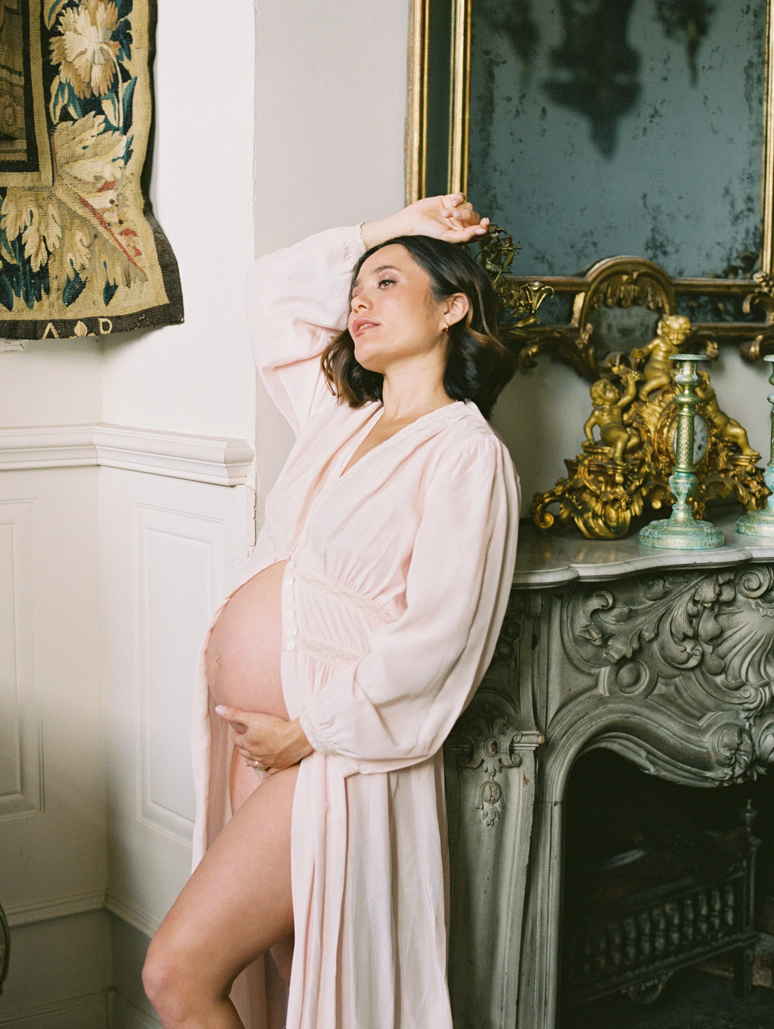 A pregnant woman wearing a pale pink robe stands in front of a stone fireplace. One arm is draped above her head, her leg is bent and her round stomach​ is exposed​. On the mantelpiece are gold French antiques.