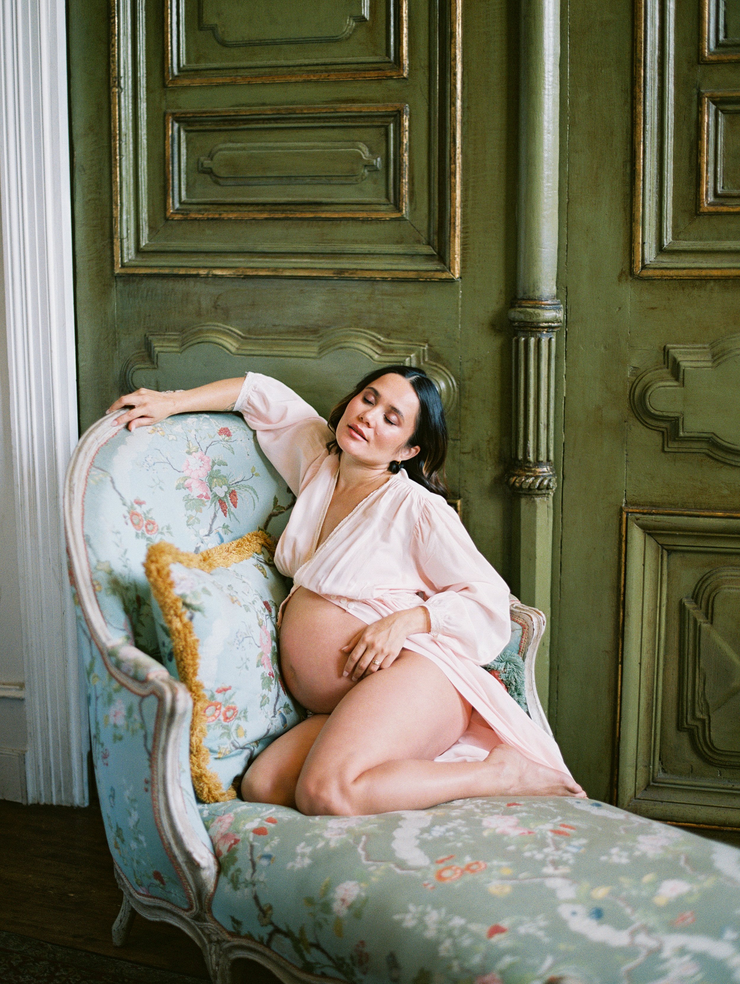A pregnant woman poses on a​n antique​ French chaise. She wears a pale pink robe ​and her round stomach​ is exposed​. Her eyes are closed. Behind her is a green​, antique do​or.