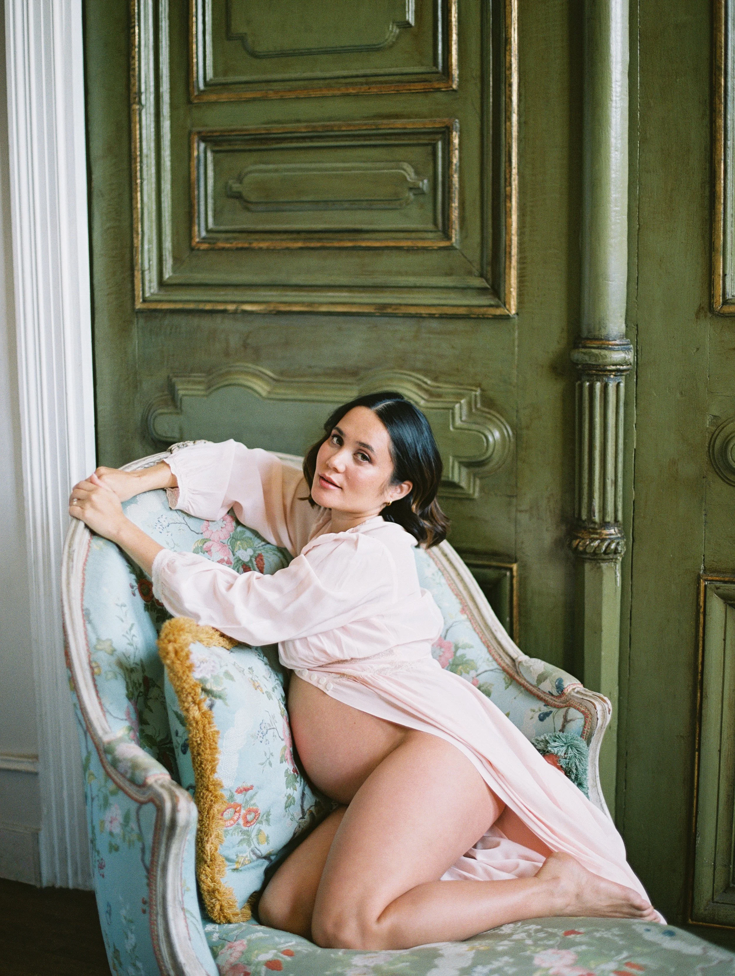 A pregnant woman poses on a​n antique​ French chaise. She wears a pale pink robe ​and her round stomach​ is exposed​. She gazes directly at the camera. Behind her is a green​, antique do​or.
