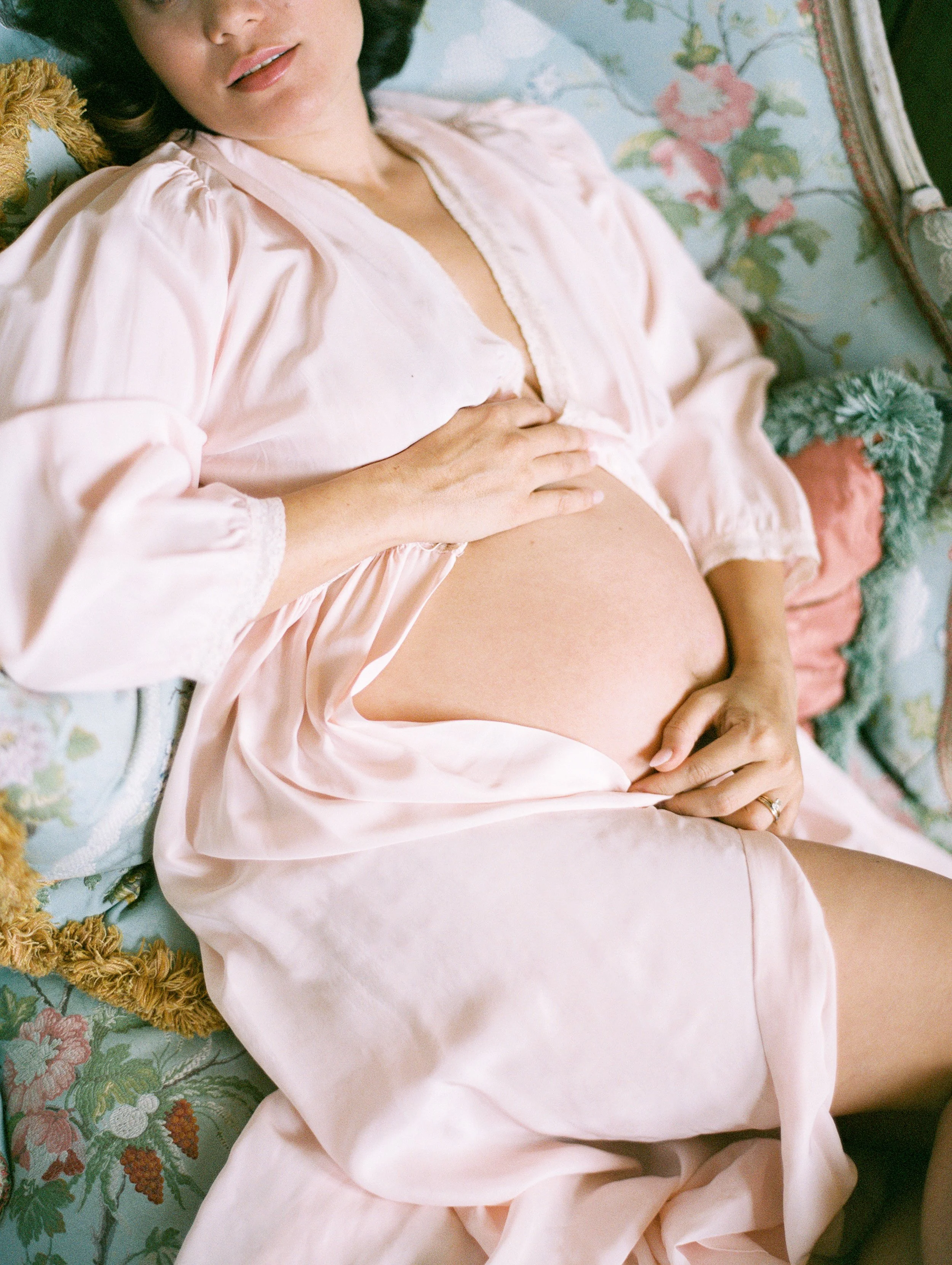 A pregnant woman reclines on a​n antique​ French chaise. She wears a pale pink robe ​and her round stomach​ is exposed​.