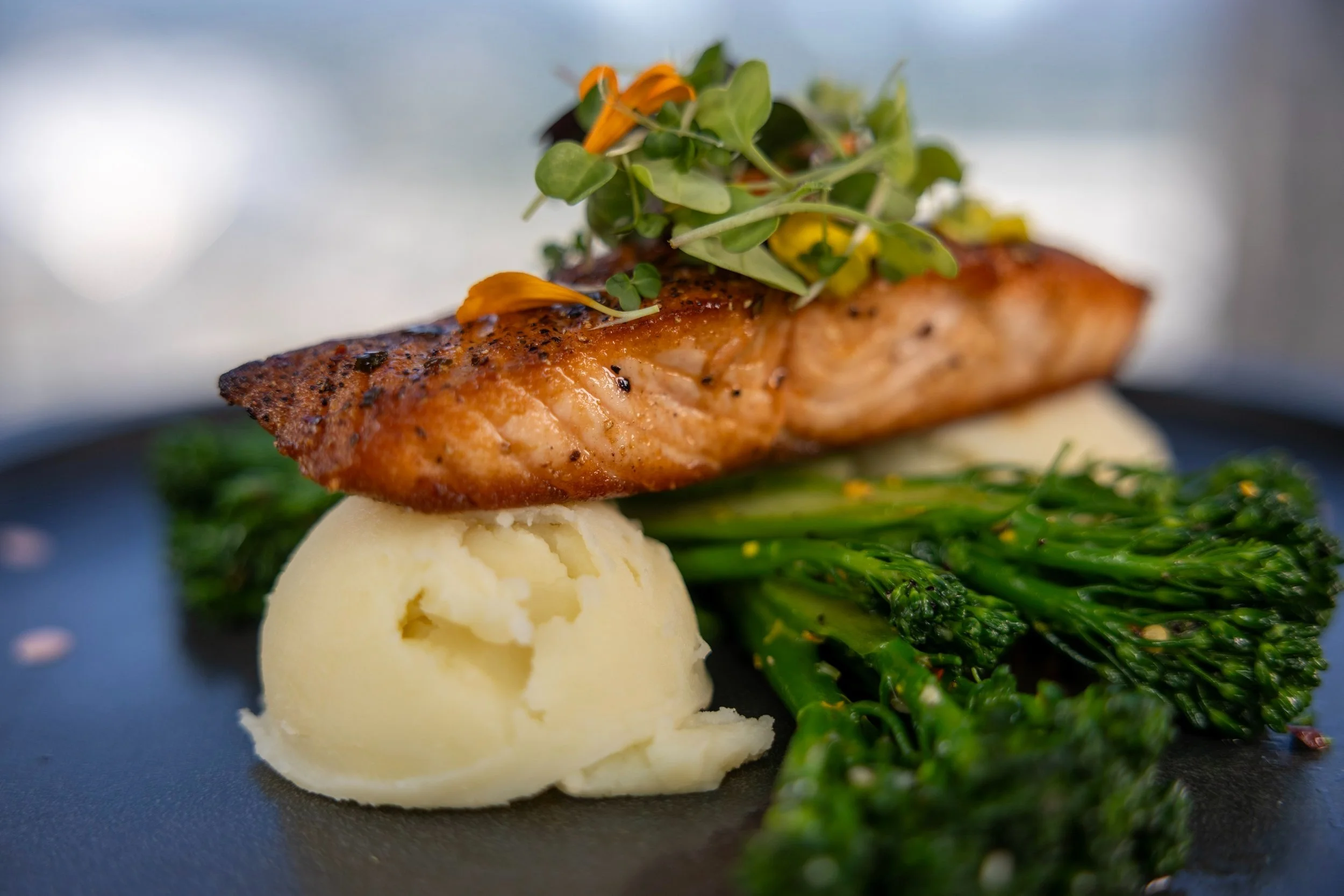 Grilled salmon fillet topped with microgreens, served with mashed potatoes and sautéed green vegetables on a black plate.