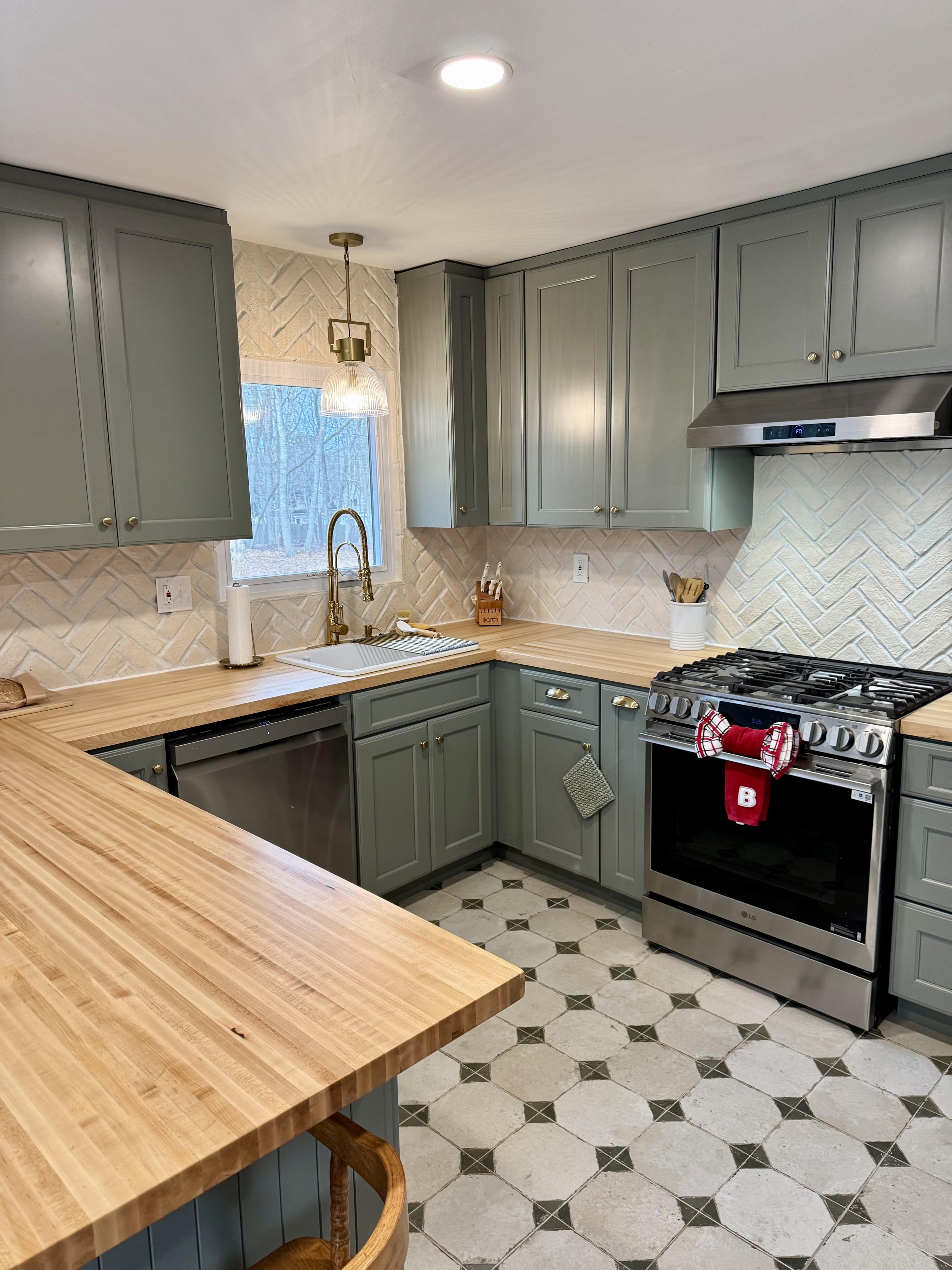 Modern kitchen with gray cabinets, wooden countertops, stainless steel appliances, a window above the sink, and patterned tile flooring.