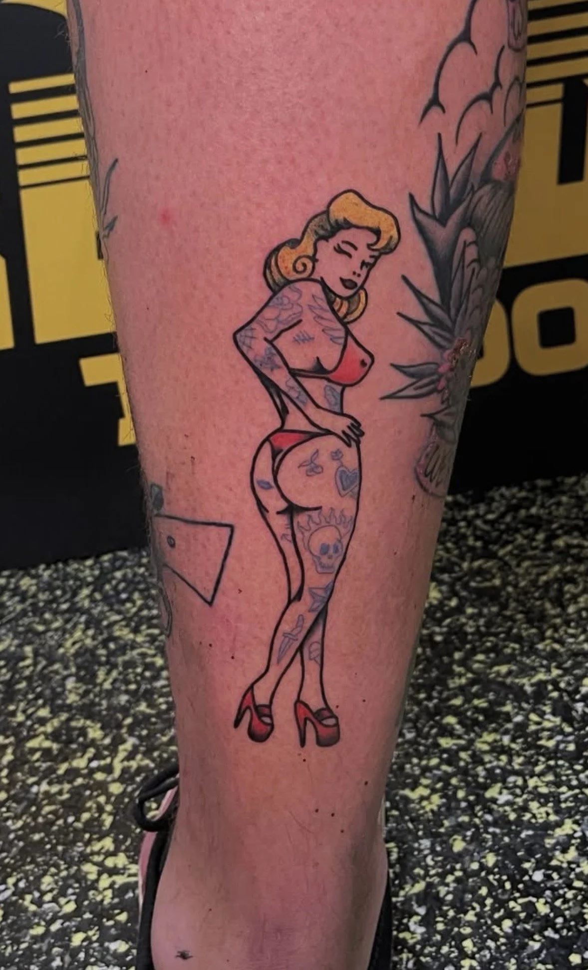 Tattoo of a cartoon woman with tattoos, wearing red underwear and high heels, standing with an arched back and face tilting downward.