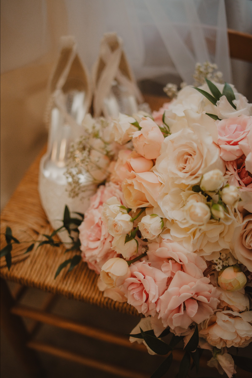 TheBloomeryKC.com. A bouquet of light pink and white roses and peonies on a wicker chair, with ballet slippers hanging in the background.
