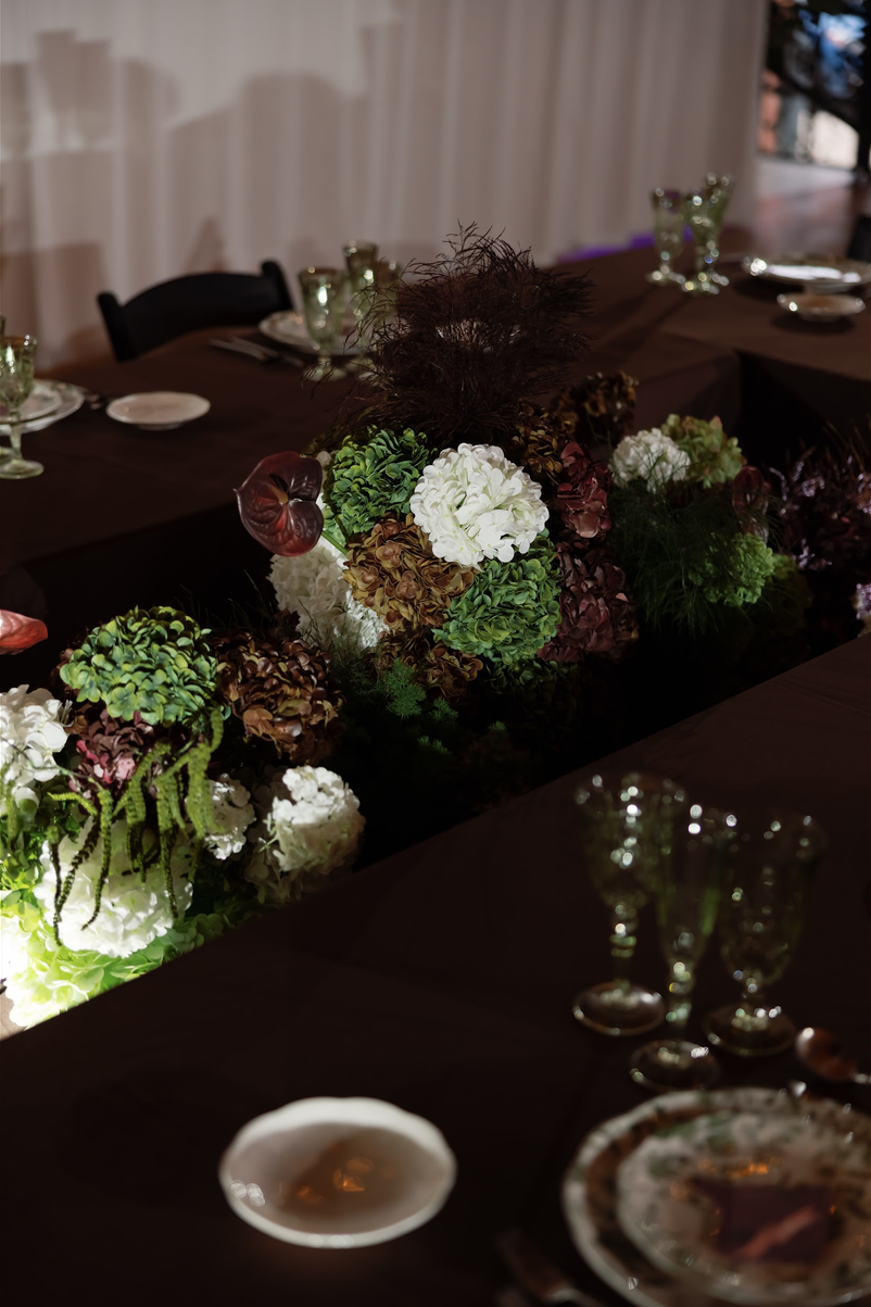 TheBloomeryKC.com. A dark dining table decorated with a floral centerpiece of white, green, and brown flowers and greenery, with glassware and plates set around it.
