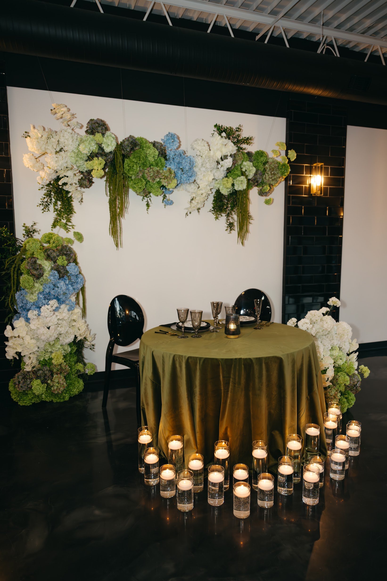 TheBloomeryKC.com. A table with a gold tablecloth, dark glasses, and a candle. There is a floral arrangement with white, green, purple, and blue flowers hanging from the wall, and candles in glass holders are placed around the table on the floor.