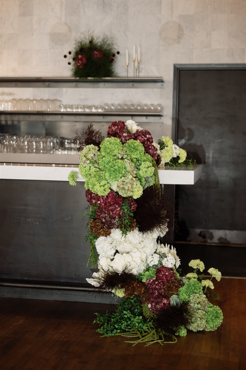 TheBloomeryKC.com. A floral arrangement with green, white, and maroon flowers, placed on a white surface in a modern, minimalistic room with glasses and a vase in the background.