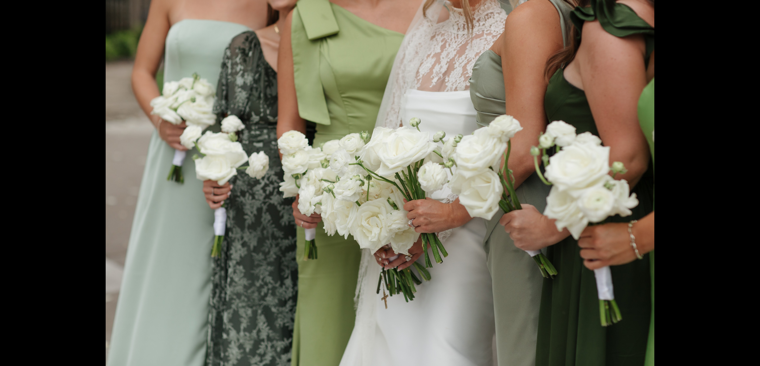 TheBloomeryKC.com. Close-up of women holding white floral bouquets at a wedding, with glimpse of bride in white gown and bridesmaids in various shades of green dresses.