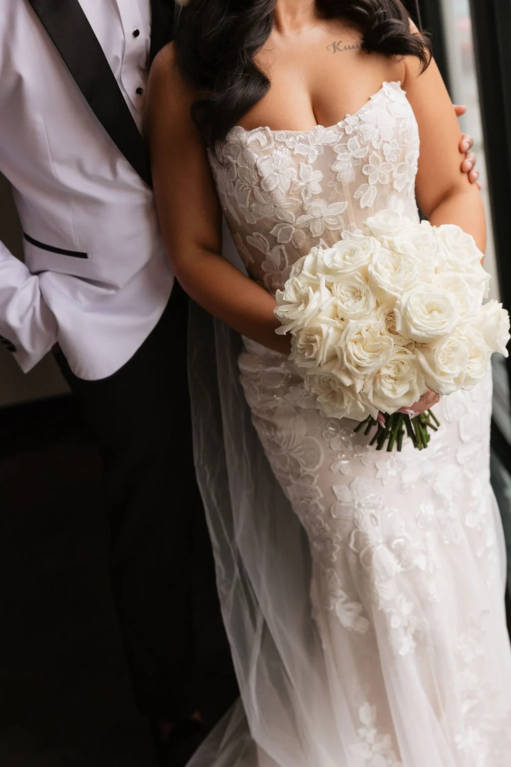 TheBloomeryKC.com. Bride holding a bouquet of white roses, dressed in a lace wedding gown, standing close to a man in a white tuxedo.