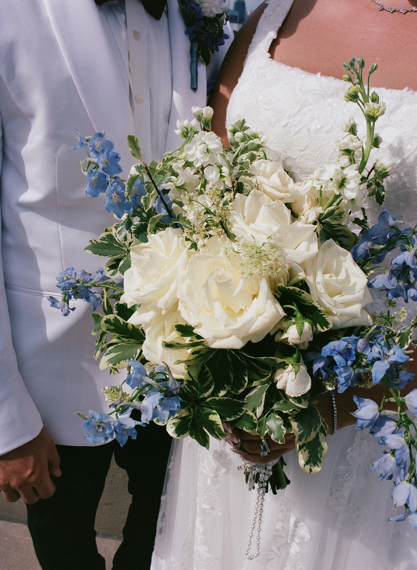 TheBloomeryKC.com. Close-up of a bride holding a bouquet of white roses, blue delphiniums, and green leaves, with a bridesmaid or groom standing nearby in a white suit and tie.