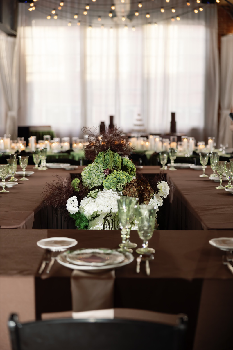 TheBloomeryKC.com. Elegant banquet table setup with a floral centerpiece featuring white and green flowers, surrounded by glassware and plates in a well-lit room with large windows and string lights.