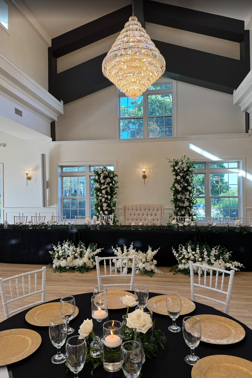 TheBloomeryKC.com. Elegant wedding reception setup with a round table in the foreground, decorated with white roses and candles, and a head table in the background adorned with white floral arrangements, and two tall floral stands.