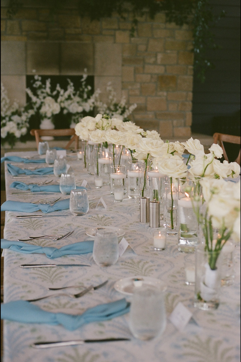 TheBloomeryKC.com. A decorated dining table with white flowers in vases, candles, and light blue napkins in a cozy indoor setting with brick walls.