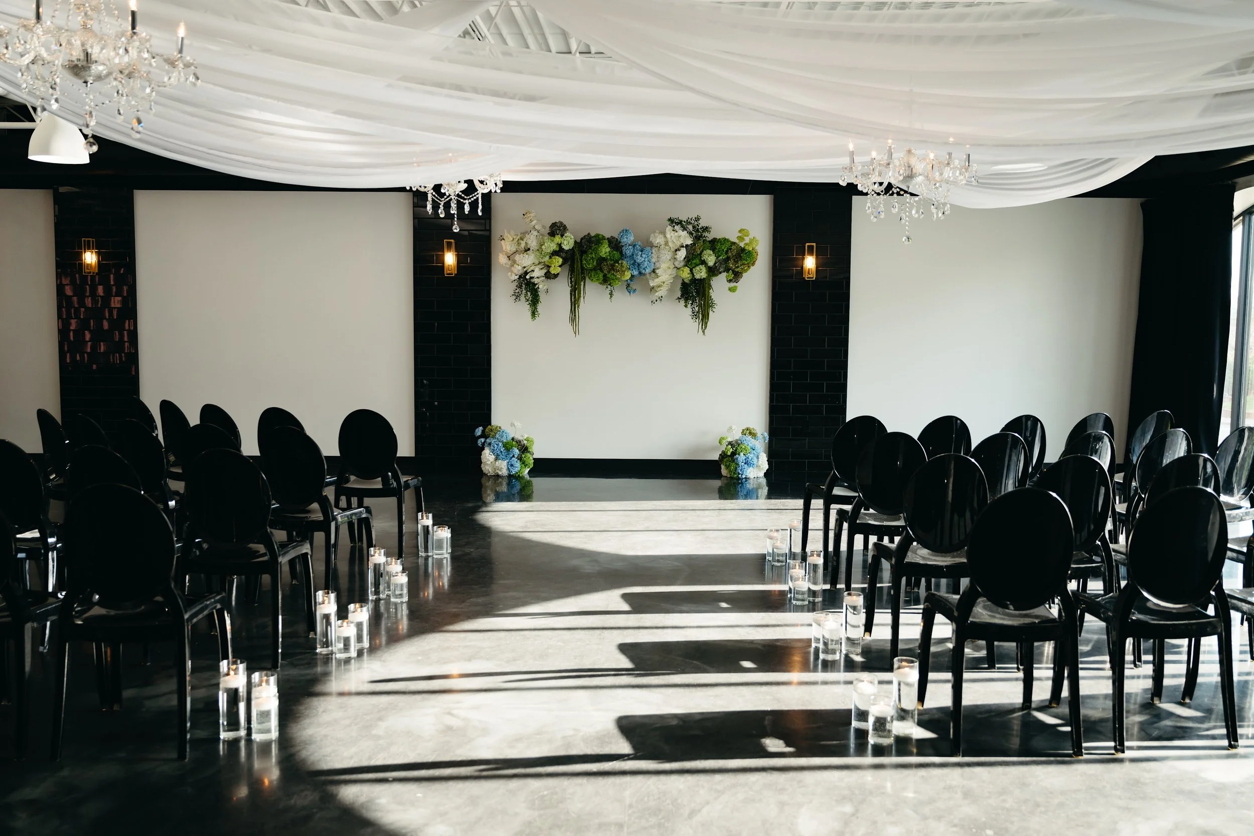 TheBloomeryKC.com. An indoor wedding ceremony setup with black chairs arranged on both sides of a central aisle, decorated with candles in glass holders, a white draped ceiling, and floral arrangements on a white backdrop with black accents.