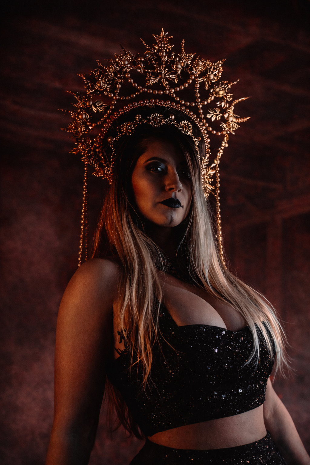 A woman with long blonde hair and dark makeup, including black lipstick and smokey eyes, wearing a black, sparkly, sleeveless crop top and matching skirt. She has an elaborate golden headdress with intricate designs, beads, and floral elements, and is standing in a dark, red-lit room.