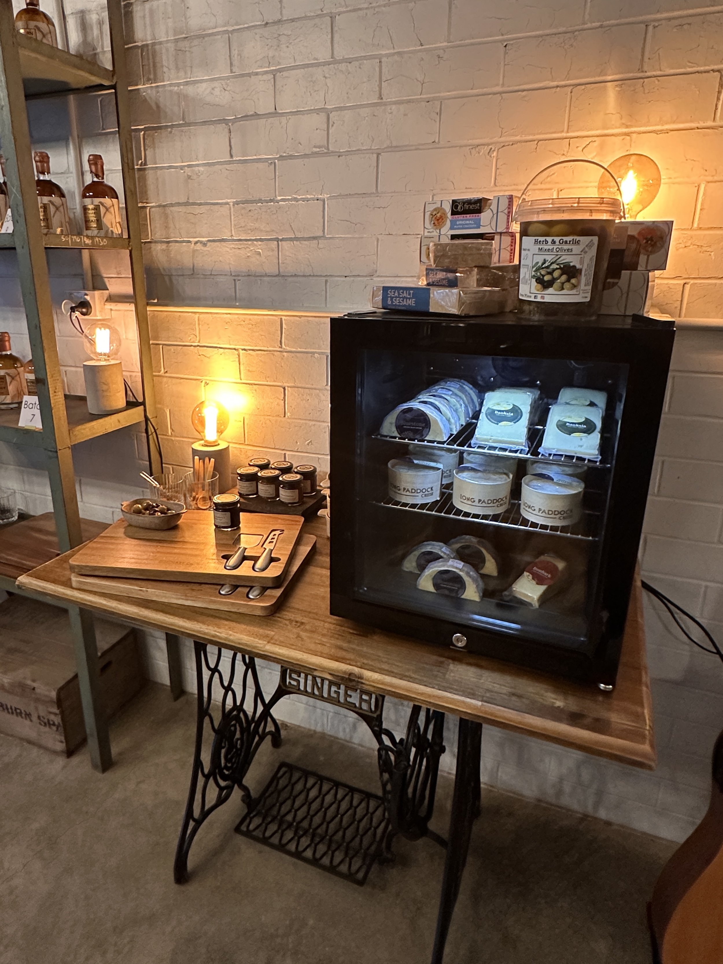 A small refrigerated display case containing cheese and dairy products, placed on a wooden table with jars of jam and snacks nearby. Shelves with bottles and boxes of food items are in the background, illuminated by warm ambient lighting.