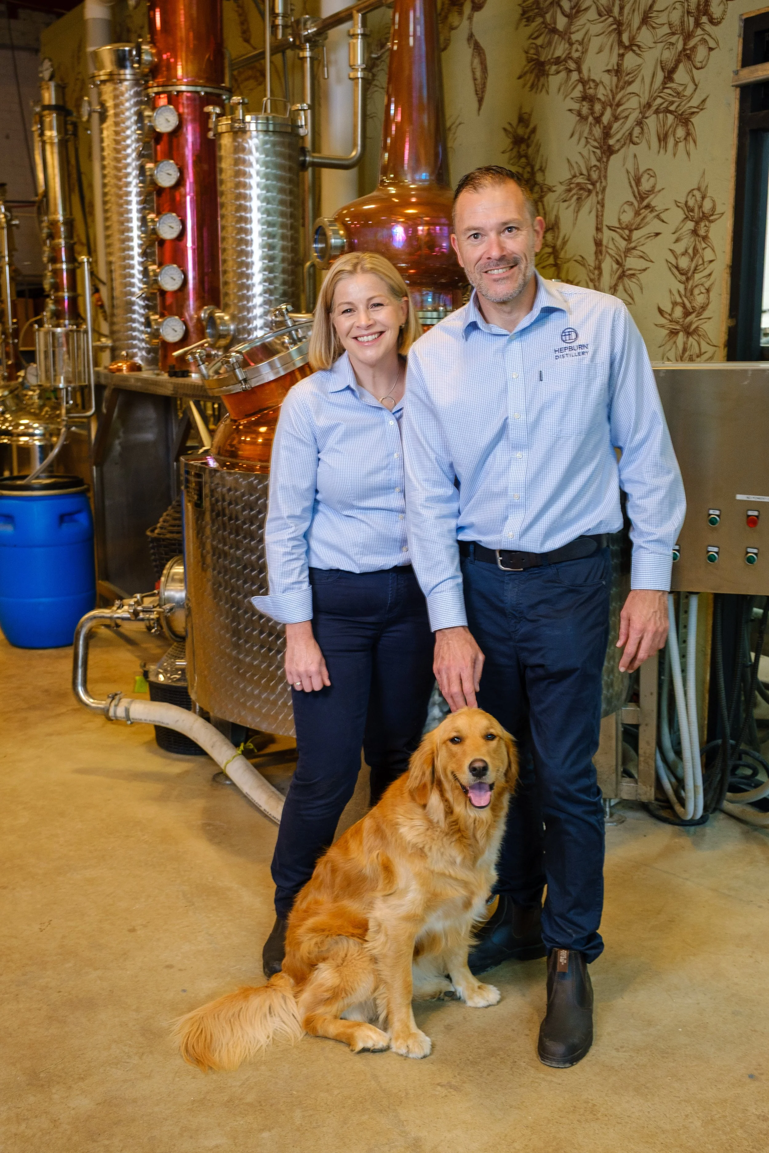 Two people, a woman and a man, standing inside a distillery with a golden retriever dog sitting in front of them. The man is petting the dog, and both are smiling. The distillery equipment has shiny metal tanks and pipes.