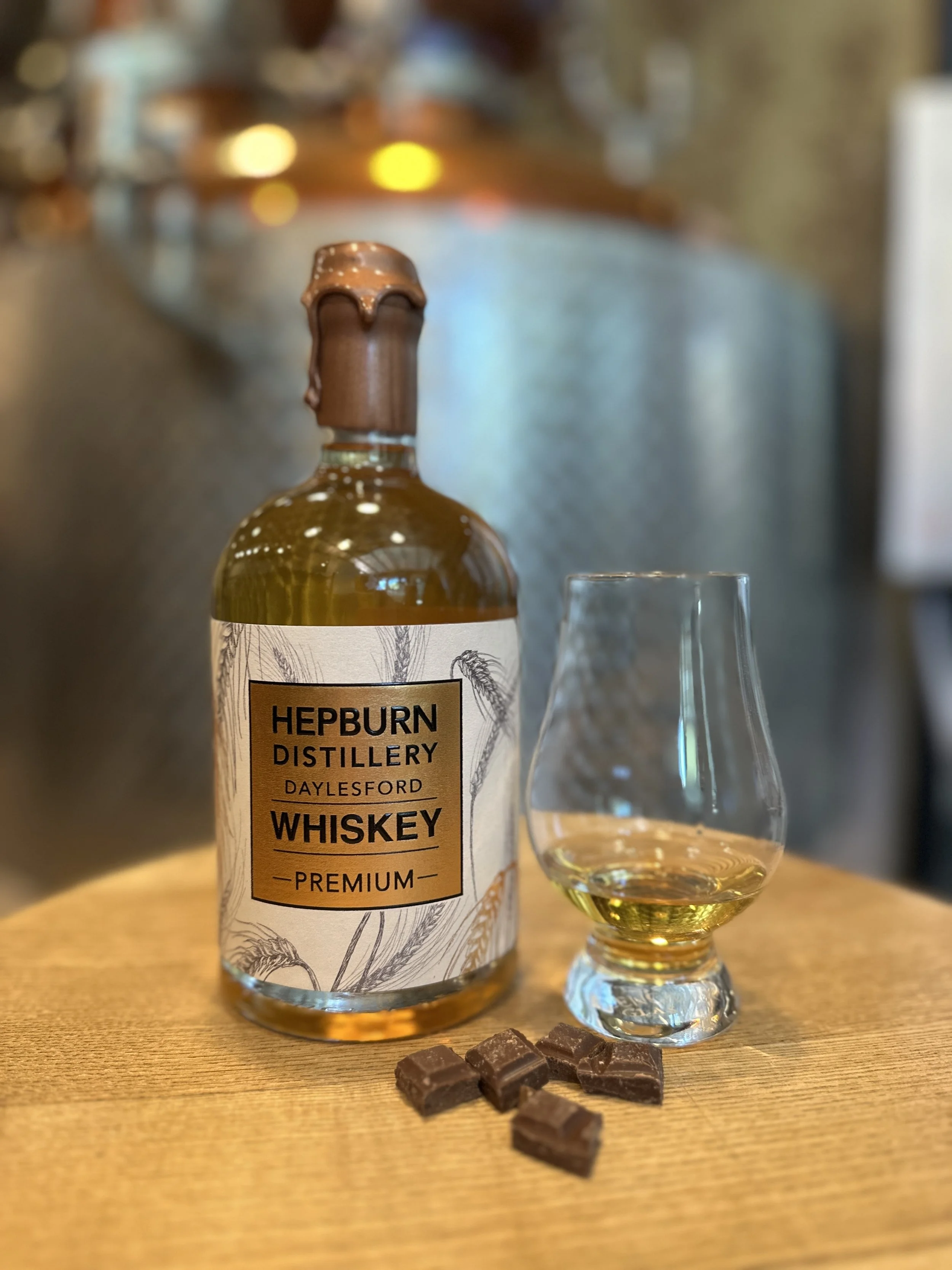 A bottle of Hepburn Distillery premium whiskey, a glass of whiskey, and pieces of chocolate on a wooden surface with a blurred background.