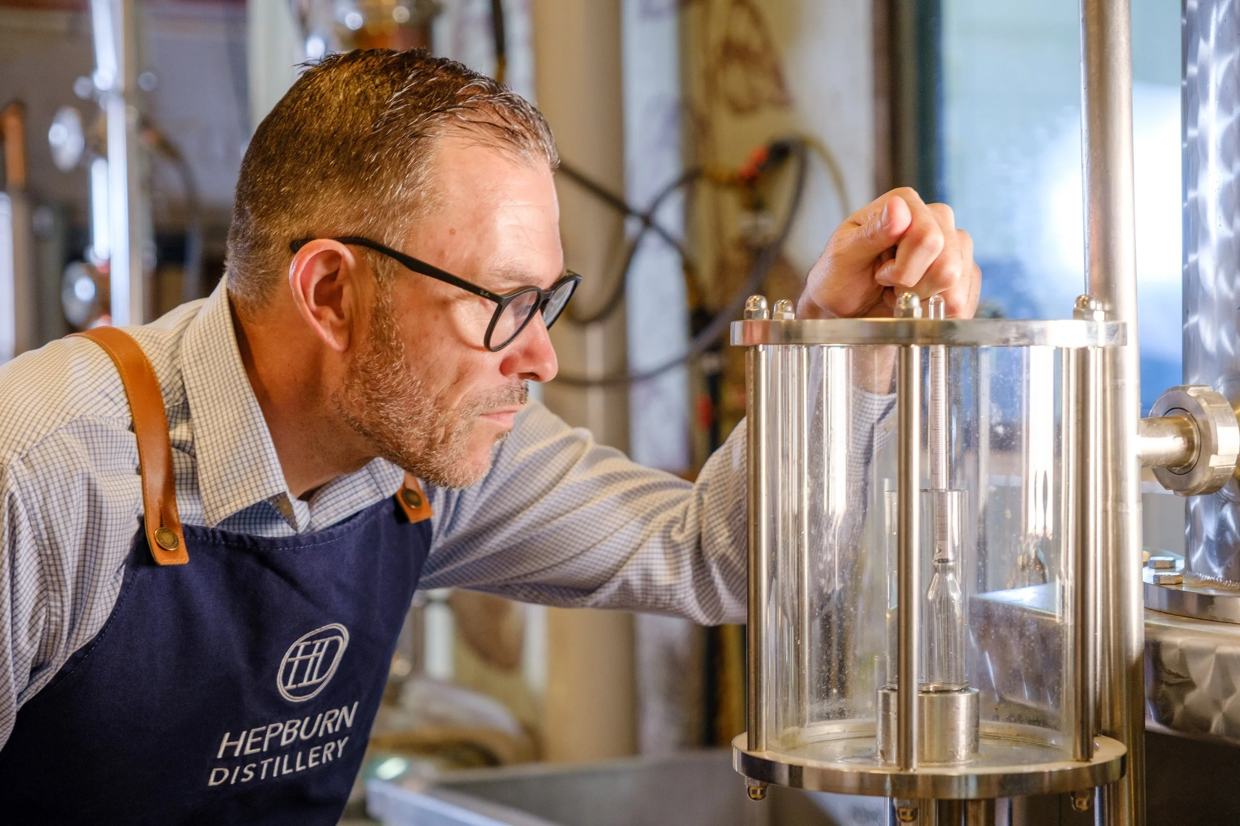 A man with glasses and a beard wearing a Hepburn Distillery apron, closely inspecting a distillation apparatus in a distillery workshop.