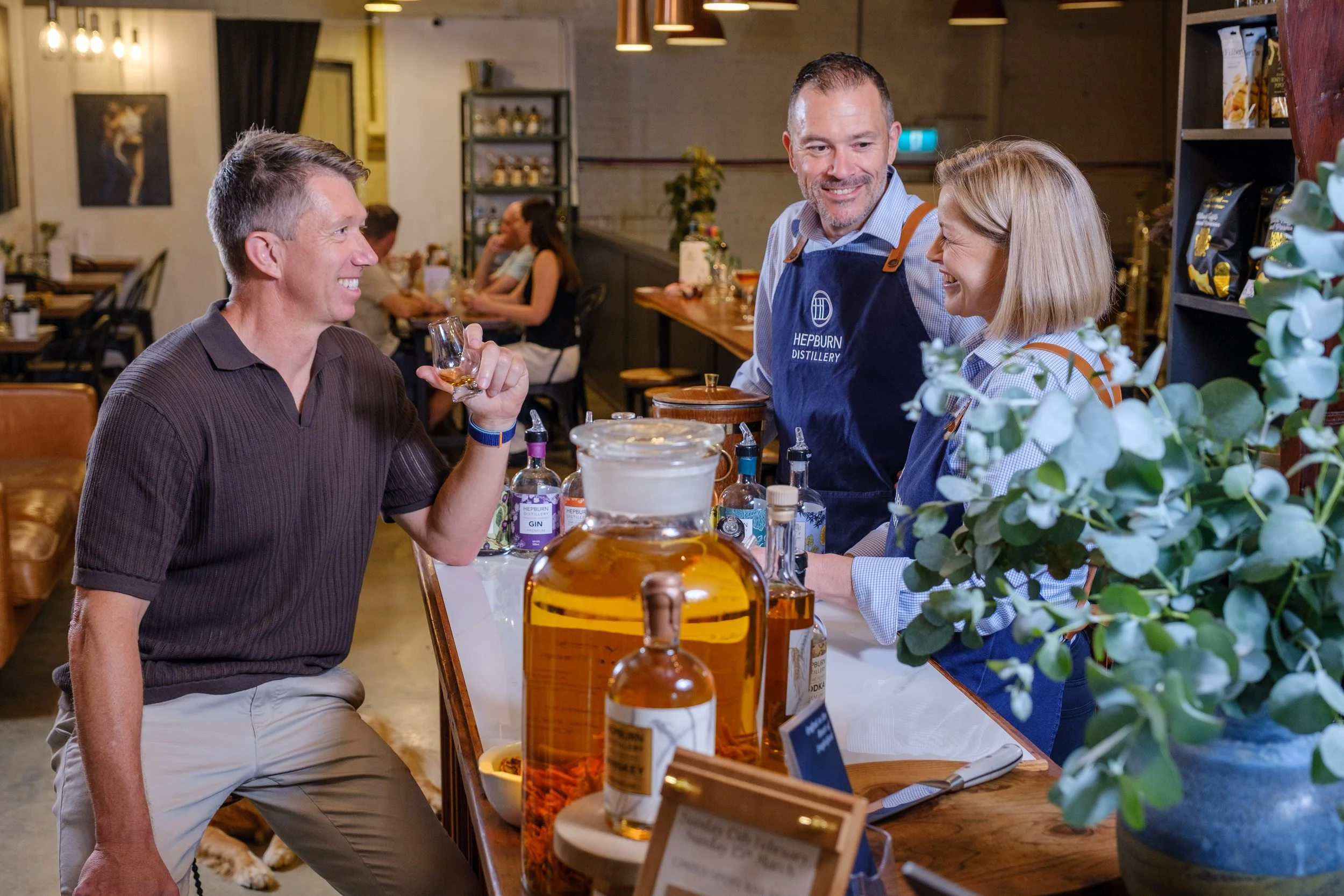 A man at a bar or tasting room pouring whiskey for a customer, with two staff members behind the counter, bottles of alcohol, and decorative plants visible.