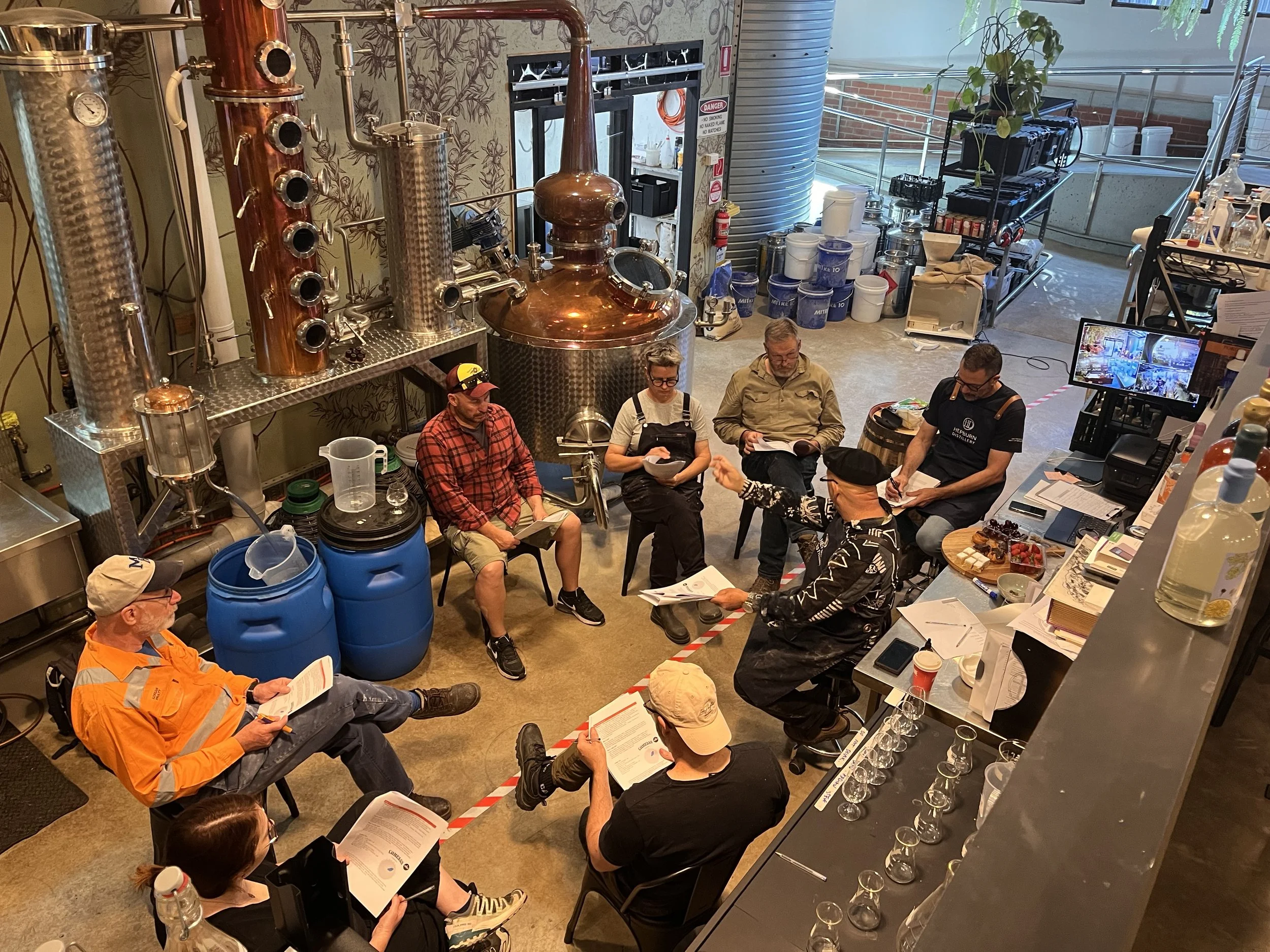 A group of people seated in a brewery, engaged in a discussion or tasting session, with brewing equipment and barrels in the background.