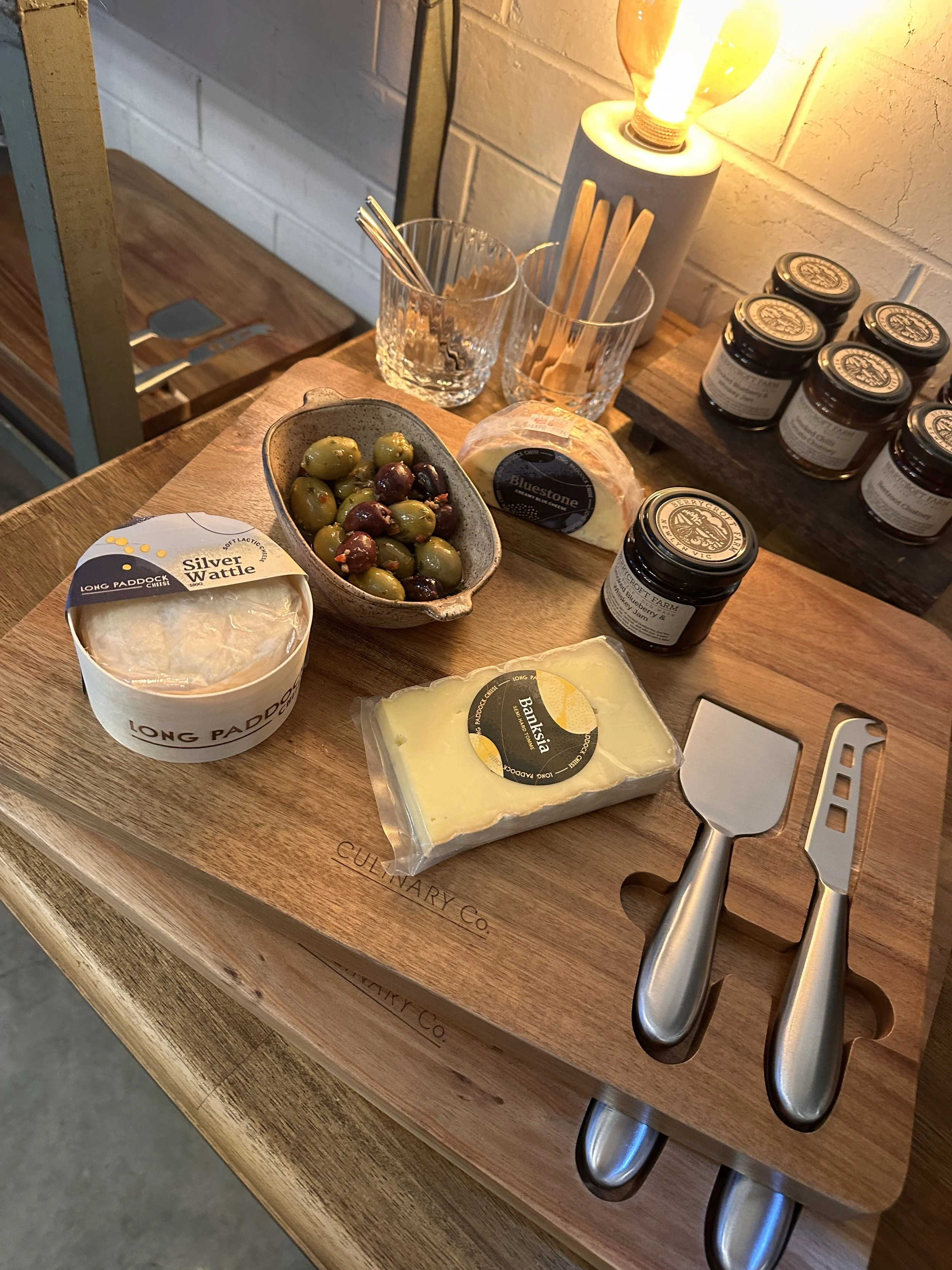 A cheese tasting board with various cheeses, a bowl of mixed green and purple olives, jars of jam, and tasting tools on a wooden tray, illuminated by a warm lamp.