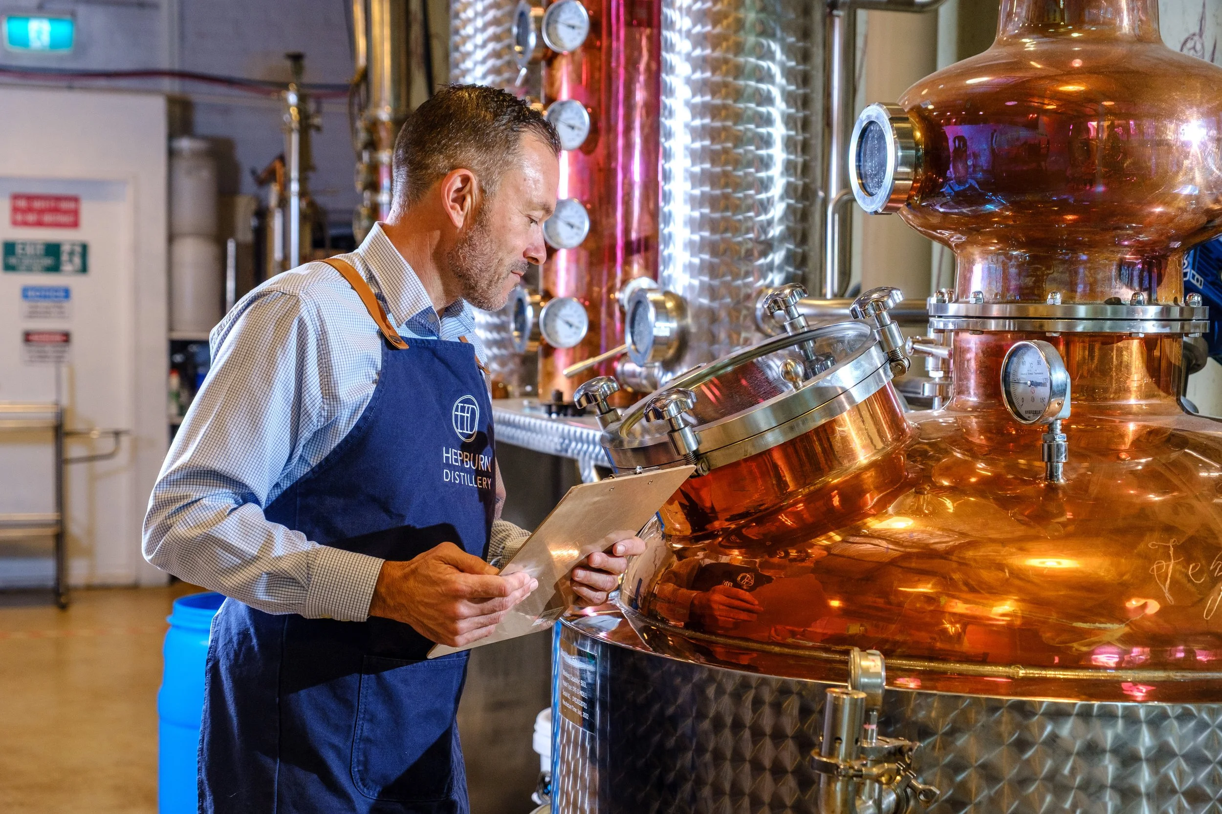 A man in a blue apron observing a large copper still at a distillery.