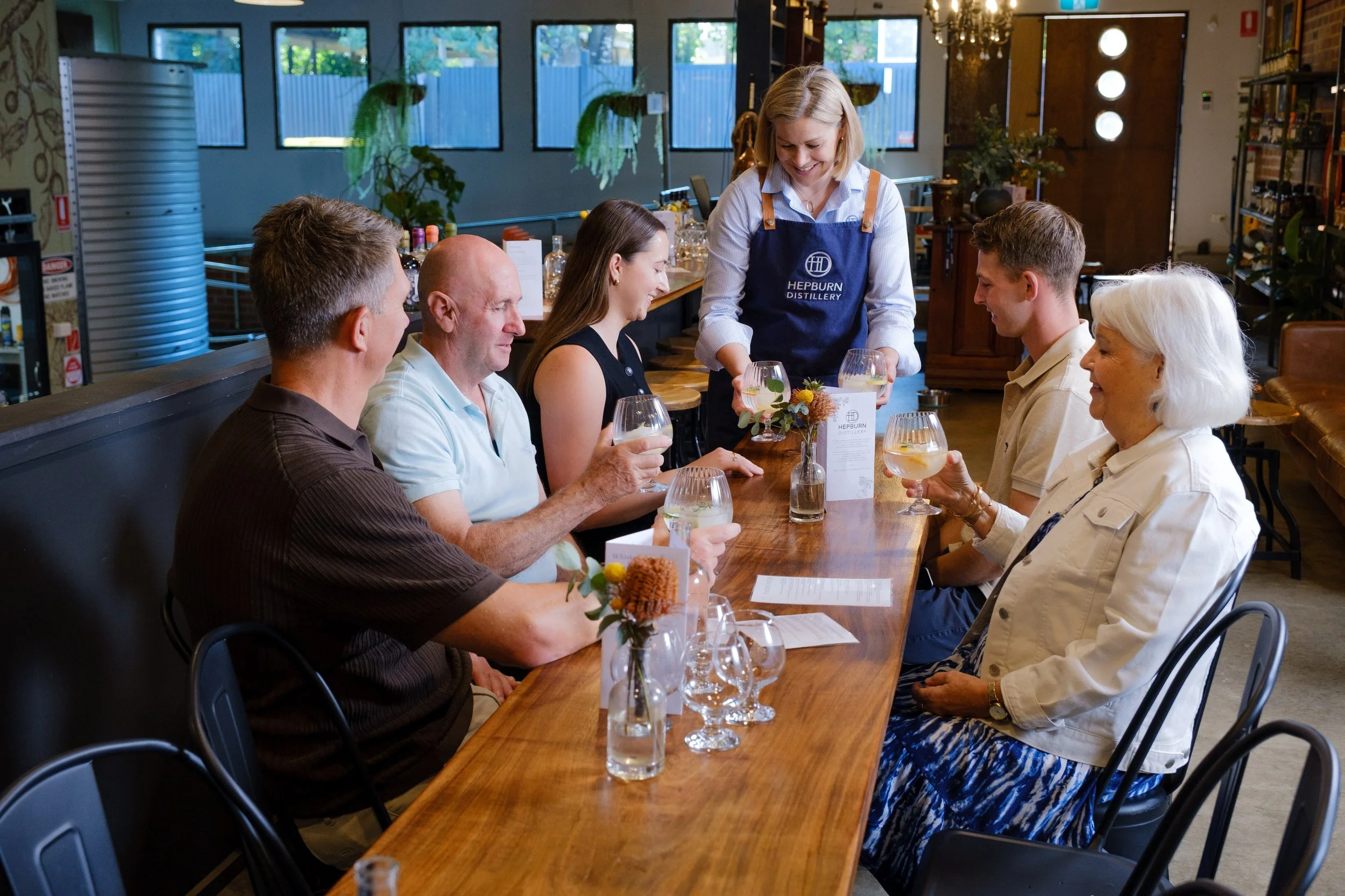 People enjoying drinks and conversation at a long wooden table in a cozy, well-lit restaurant or bar with large windows, plants, and warm decor.